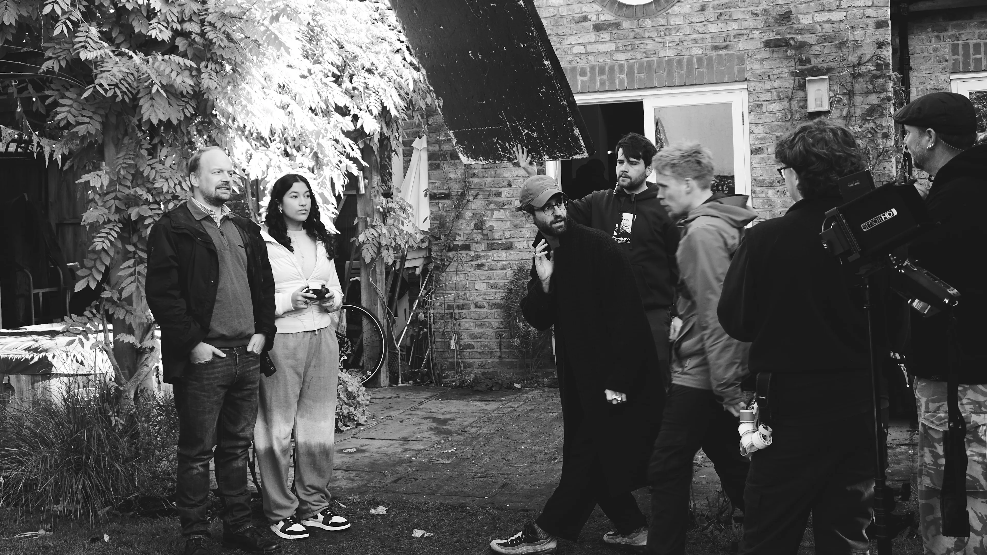A group of people, some holding film equipment, stand together in a garden area outside a brick building. The scene appears to be a film set, with casual attire and a relaxed atmosphere.