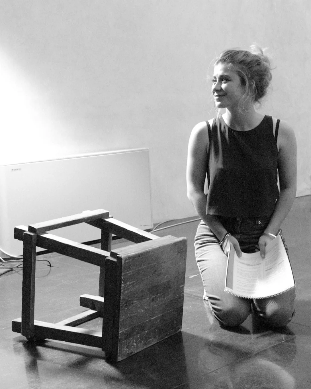 A woman kneels on the floor, smiling and holding papers, next to an overturned wooden stool. The scene is in black and white with a plain background.