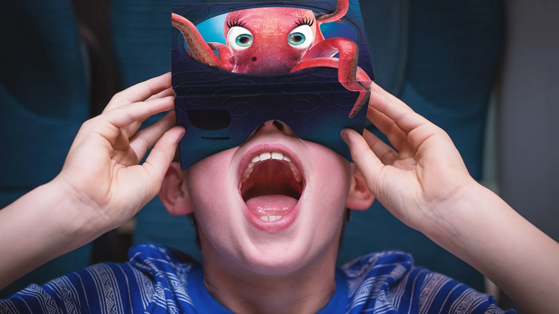 A child wearing a blue striped shirt holds a virtual reality headset with a cartoon octopus image in front of their face, mouth open wide in excitement or surprise.