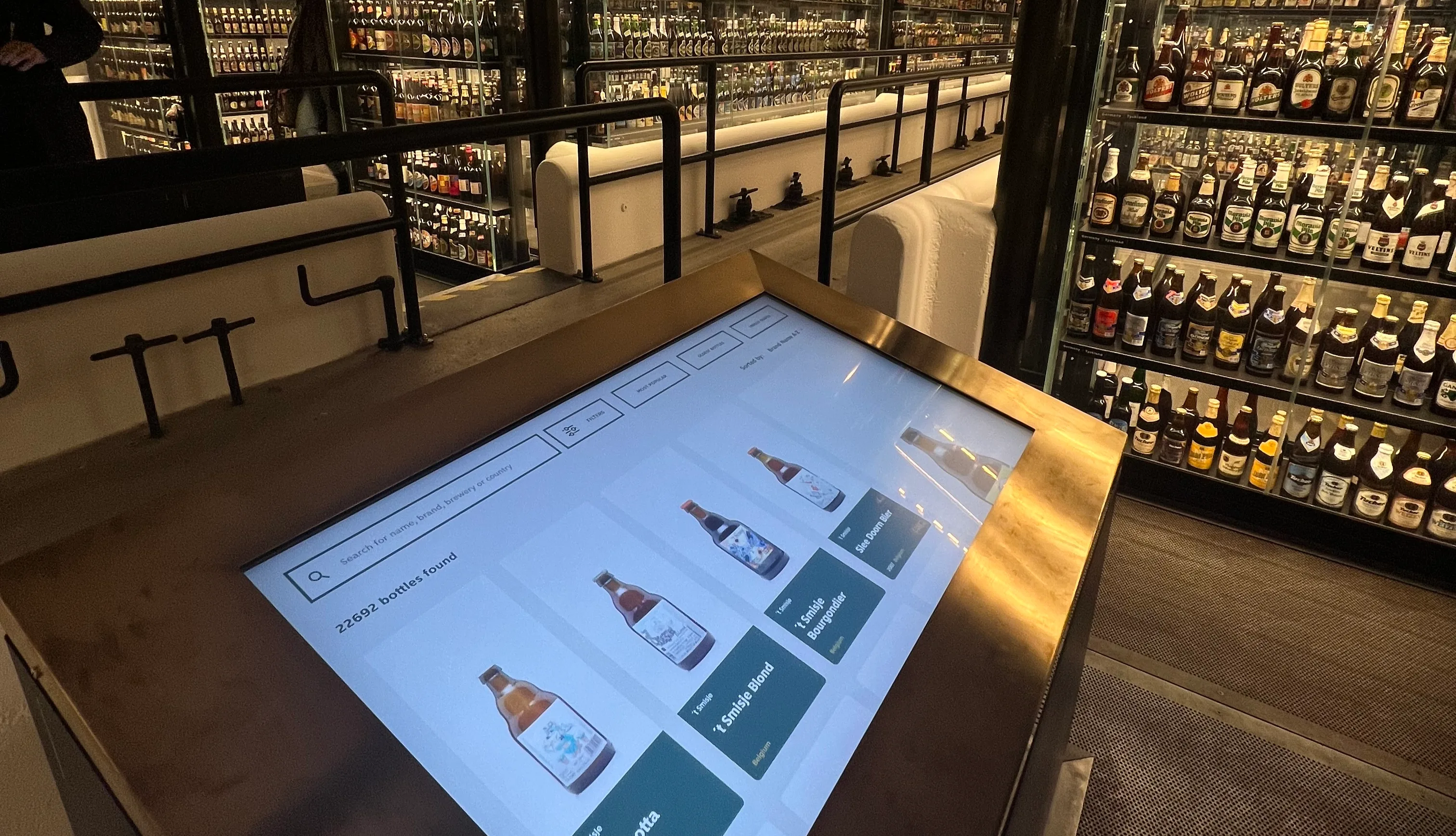 A large touchscreen display shows different bottles of whisky, with shelves lined with many more bottles of whisky in the background at a modern, well-lit liquor store or whisky museum.