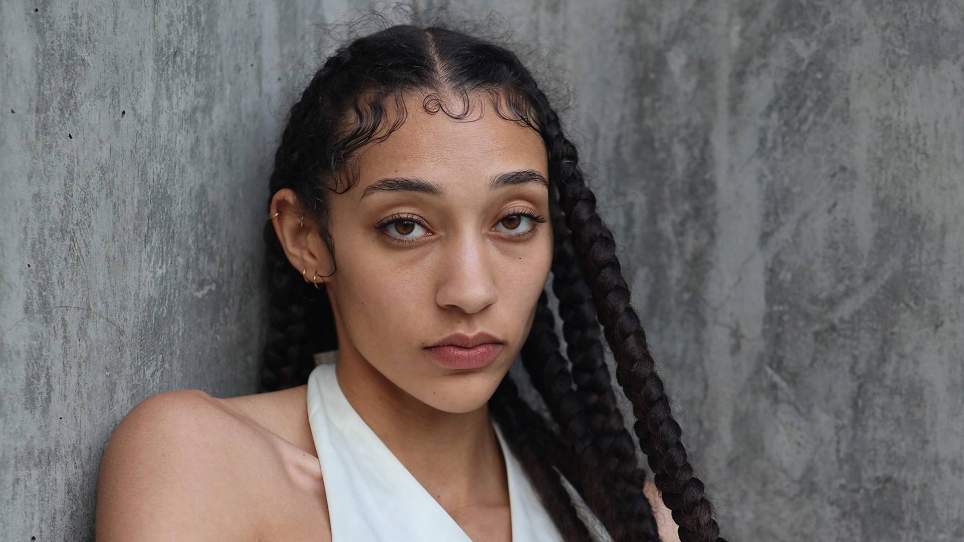 A young woman with long braids and soft curls by her forehead stands against a rough concrete wall, wearing a white halter top and gazing calmly at the camera.