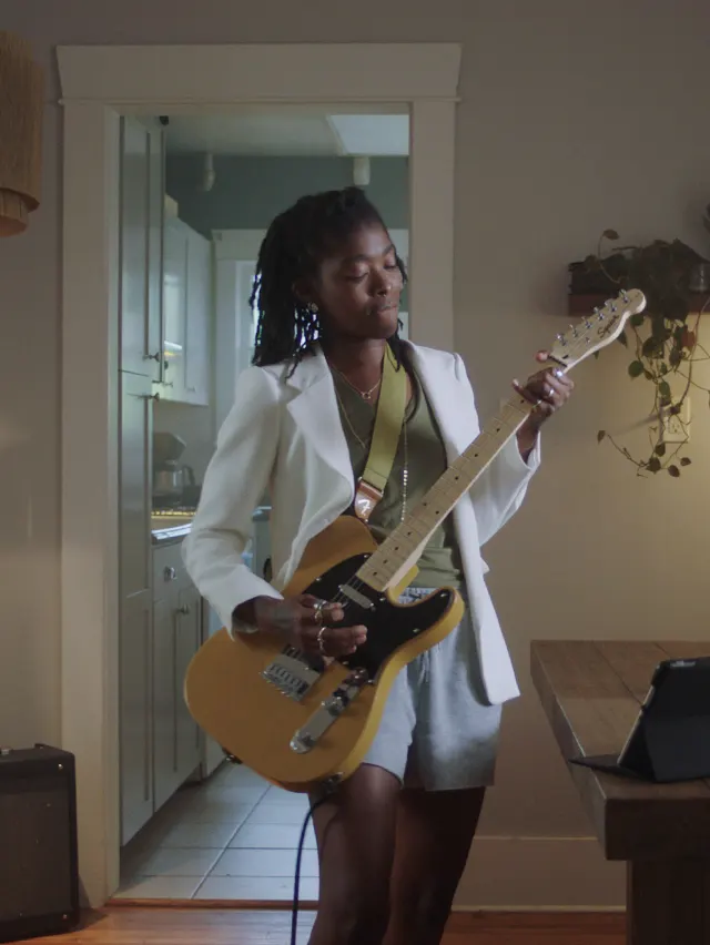 A person wearing a white blazer and gray shorts stands indoors, playing an electric guitar. There’s a wooden table, a plant, and an amplifier in the room, with a kitchen visible in the background.