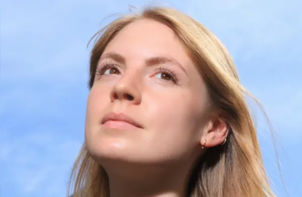 A young woman with light skin and straight, blonde hair looks upward against a bright blue sky. Sunlight highlights her face, and she wears a small earring.