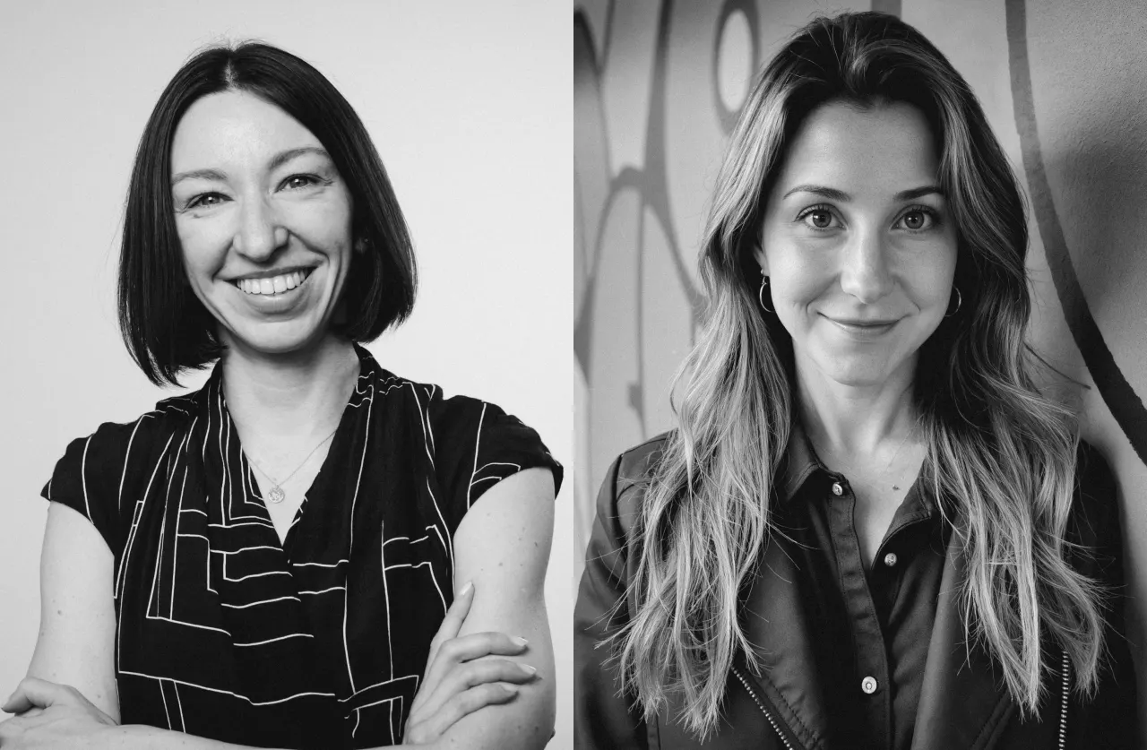 Black and white image of two women; the left woman has straight, shoulder-length dark hair, smiles broadly, and has arms crossed. The right woman has long, wavy hair and looks calmly at the camera.