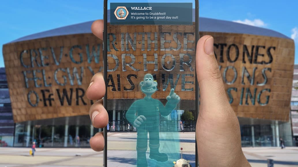 A hand holds a smartphone showing an augmented reality image of Wallace from Wallace & Gromit in front of the Wales Millennium Centre in Cardiff, with a welcome message on the screen.