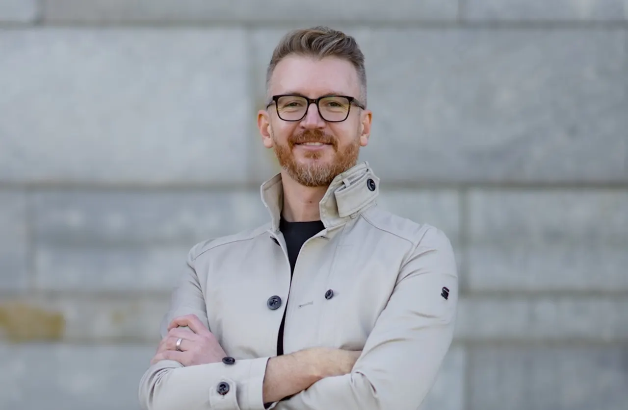 A man with short light brown hair, glasses, and a beard stands confidently with arms crossed, wearing a light-colored jacket, in front of a gray stone wall.