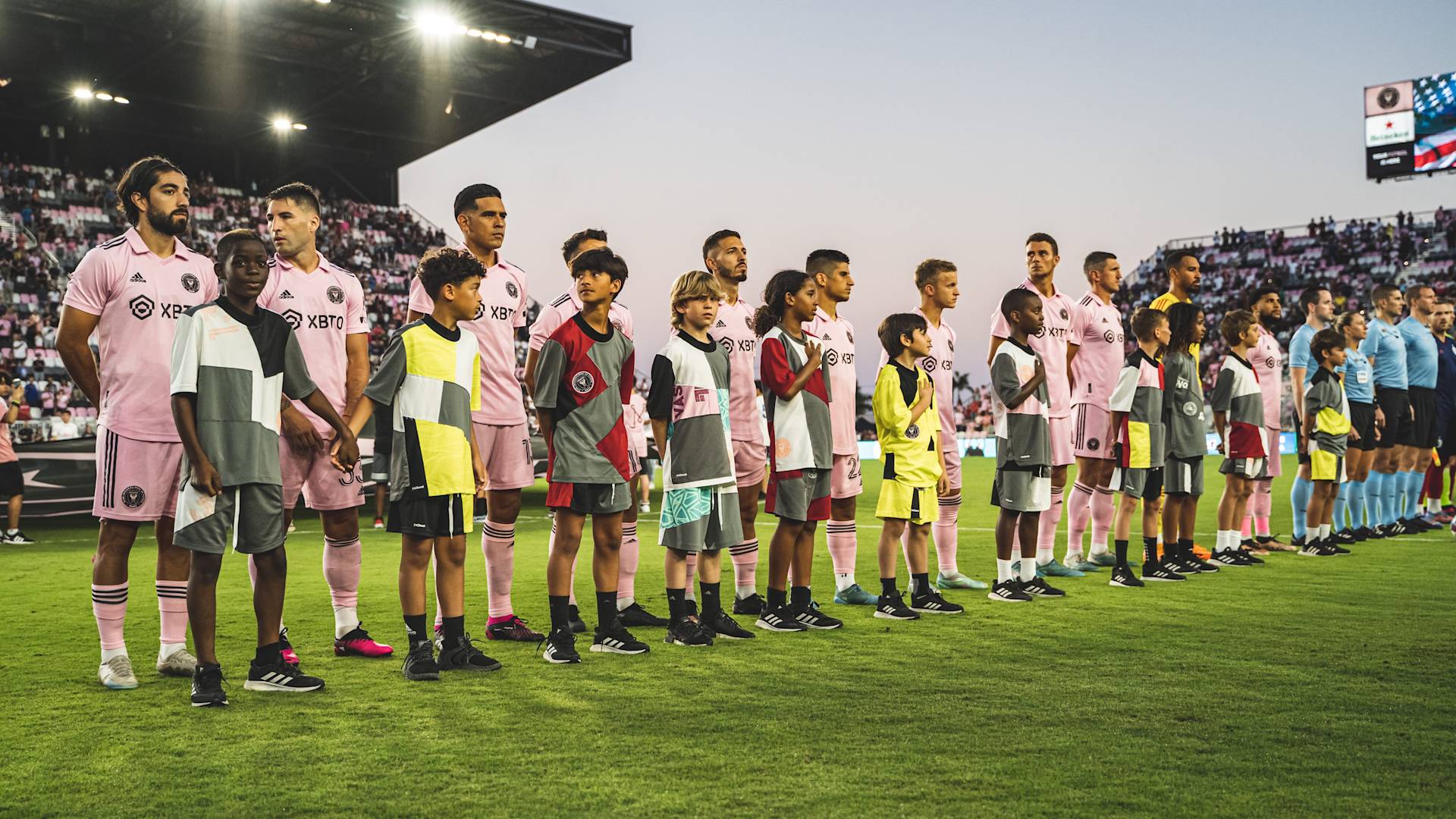 A soccer team in pink uniforms stands in a line on the field with children in front of them. Referees and a crowd of spectators are visible in the background at a stadium.