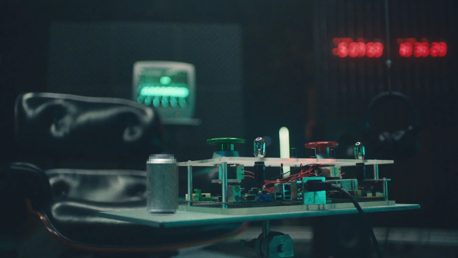 A close-up of a table with electronic components, green and red buttons, a soda can, and cables. In the background, there is a leather chair and a glowing green monitor, with red digital numbers on a wall display.