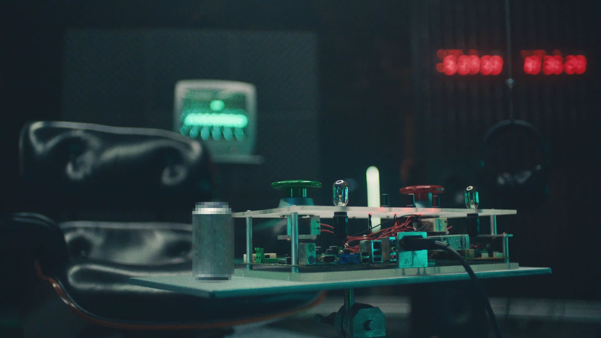 A close-up of a table with electronic components, green and red buttons, a soda can, and cables. In the background, there is a leather chair and a glowing green monitor, with red digital numbers on a wall display.