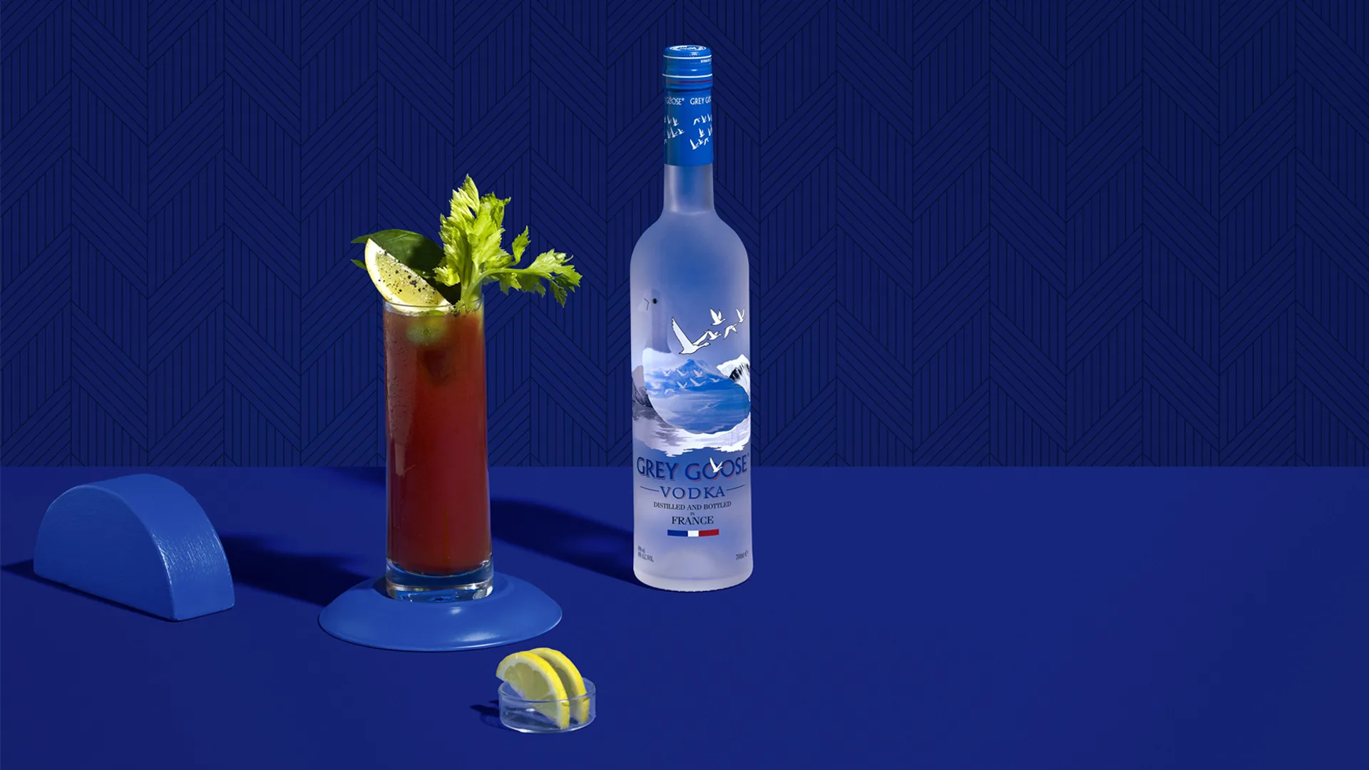 A tall Bloody Mary cocktail garnished with celery and lime sits next to a Grey Goose vodka bottle on a blue surface with a blue geometric background. A lemon wedge rests in a small container nearby.