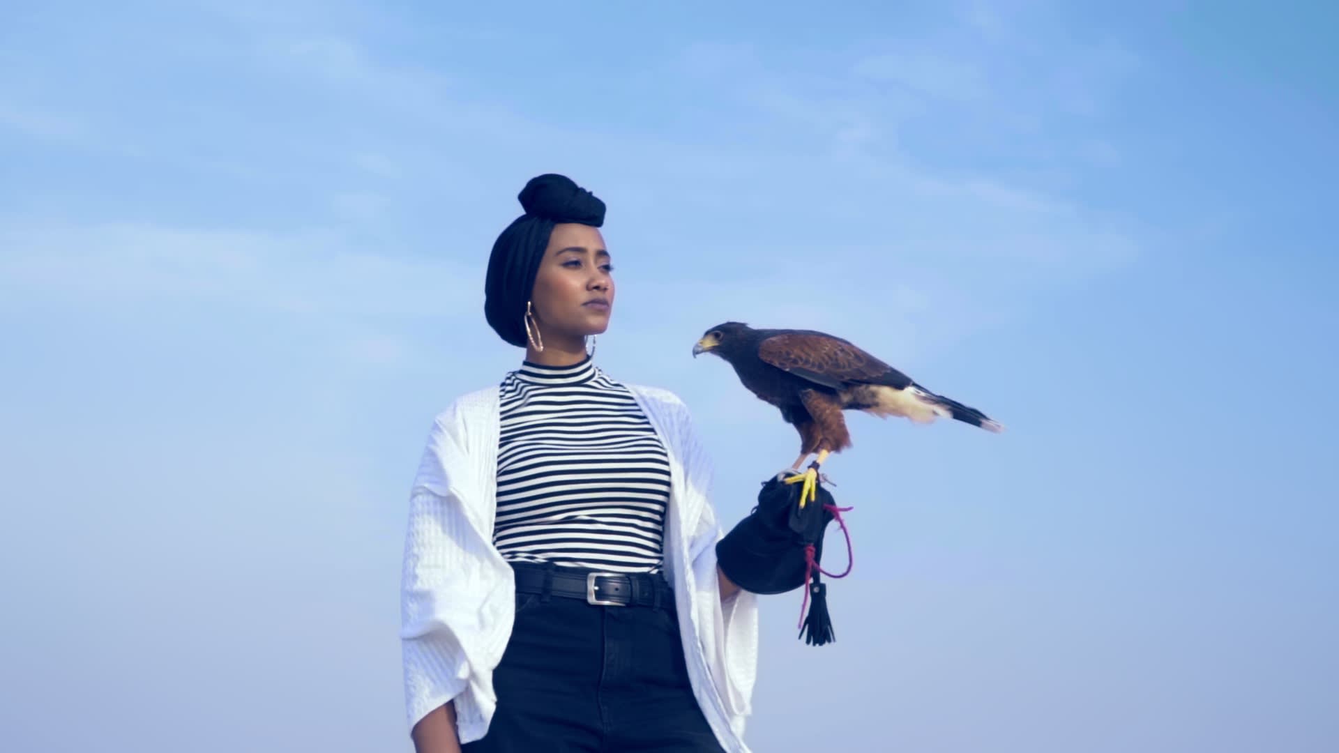 A woman in a striped shirt, white jacket, and black pants stands confidently outdoors, holding a brown hawk on her gloved arm, with a clear blue sky in the background.