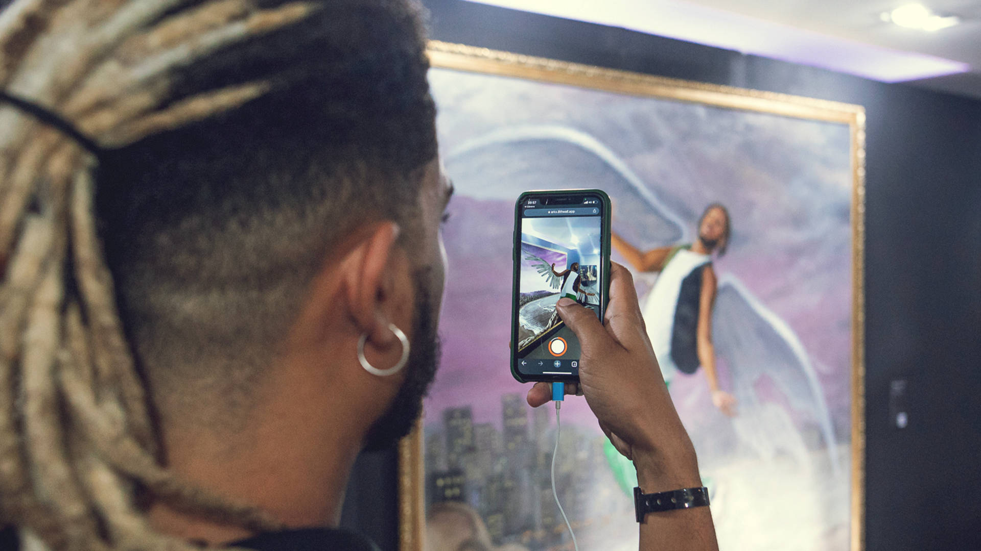 A person with light blonde dreadlocks and a hoop earring takes a photo of a large framed painting using their smartphone in an art gallery. The painting depicts a figure with outstretched arms and an urban background.