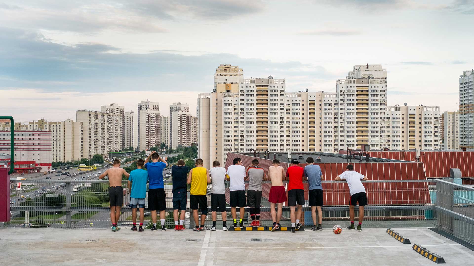 A group of eleven young people stand in a row on a rooftop, facing away and looking at a cityscape of tall apartment buildings. Soccer balls are on the ground near them. The sky is partly cloudy.