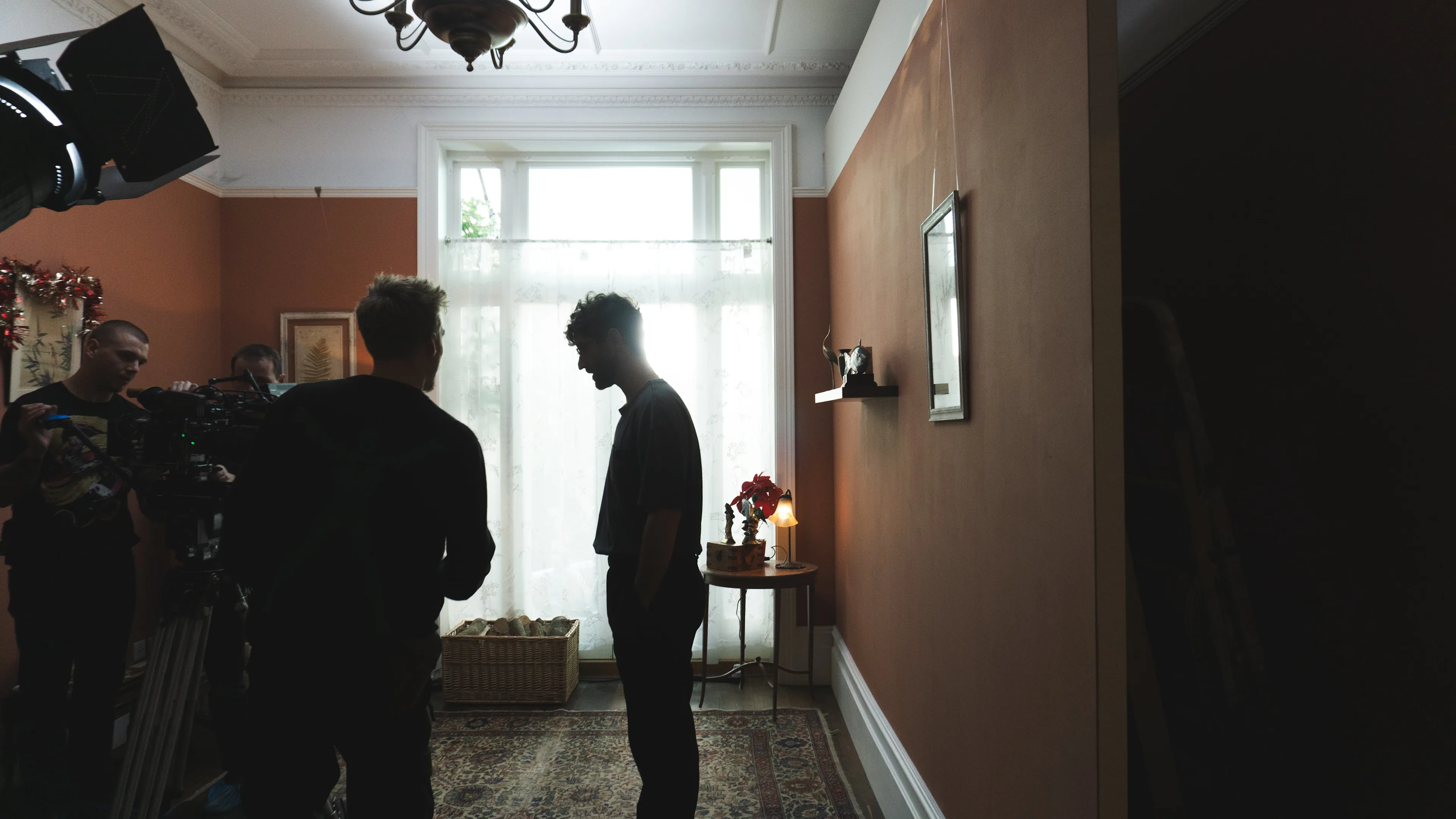 A film crew works in a dimly lit room with peach walls and a large window covered by sheer curtains. Two people stand in silhouette near the window, while others set up equipment on the left.