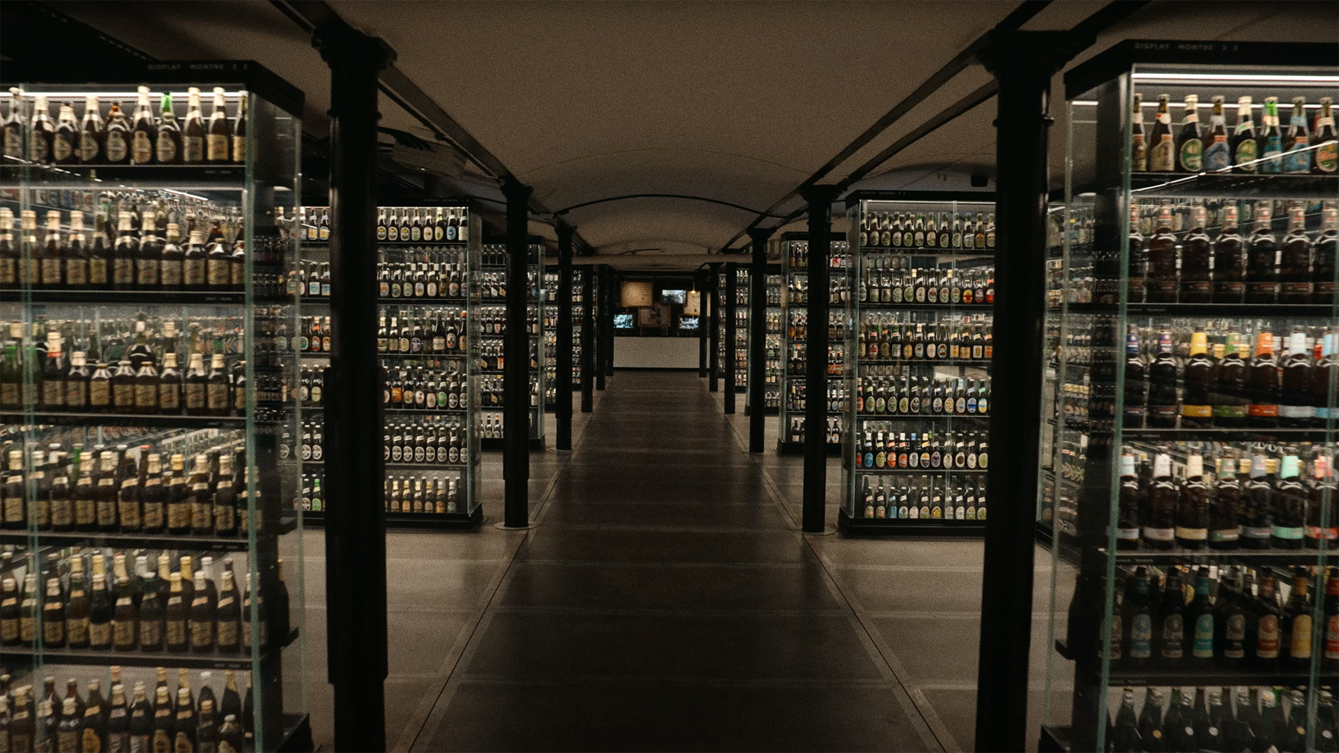Rows of glass display cases filled with various bottles line a dimly lit, arched hallway, giving the appearance of a bottle or beverage collection in a museum or exhibition setting.
