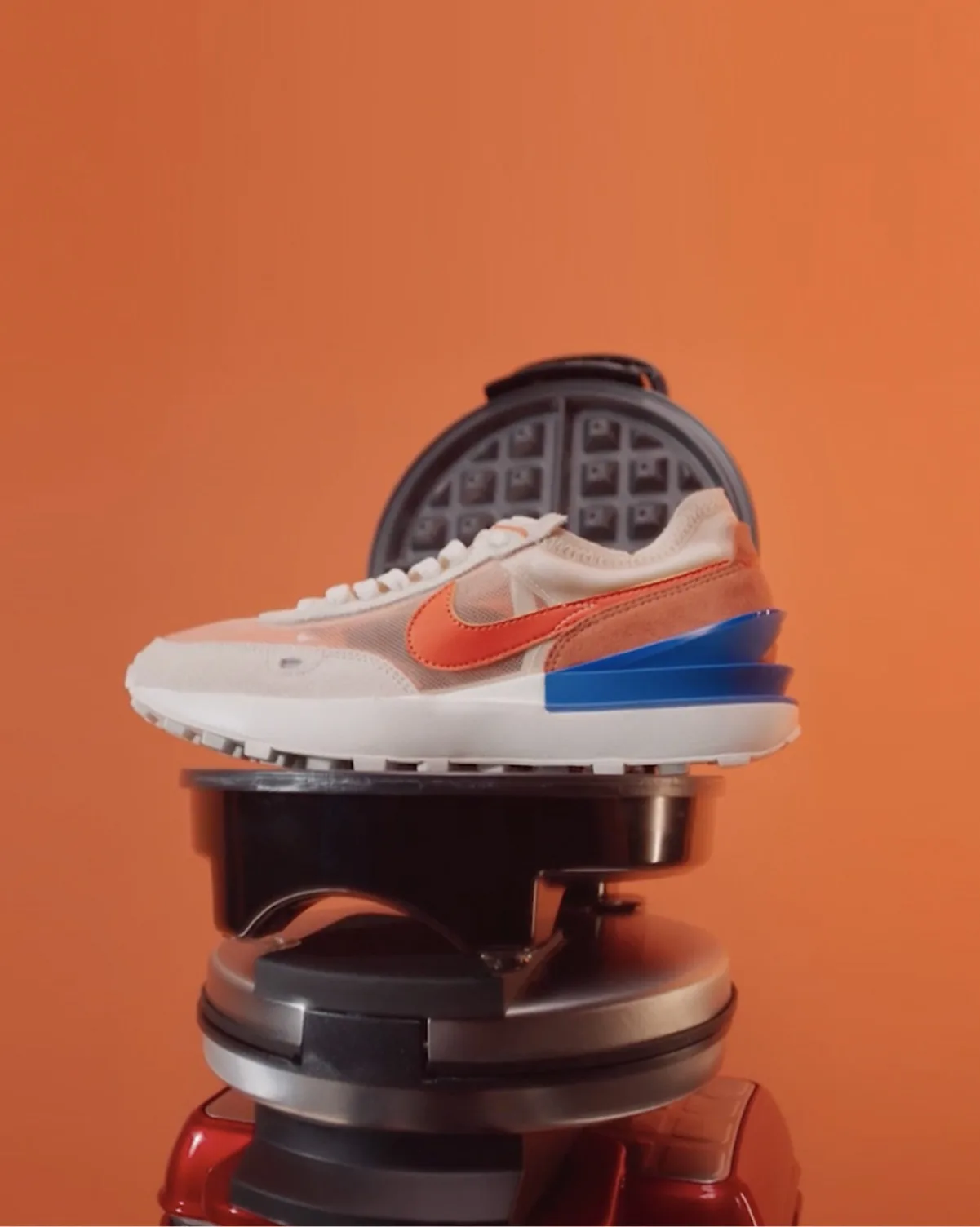 A white sneaker with orange and blue accents is placed on top of an open waffle maker, with an orange background.