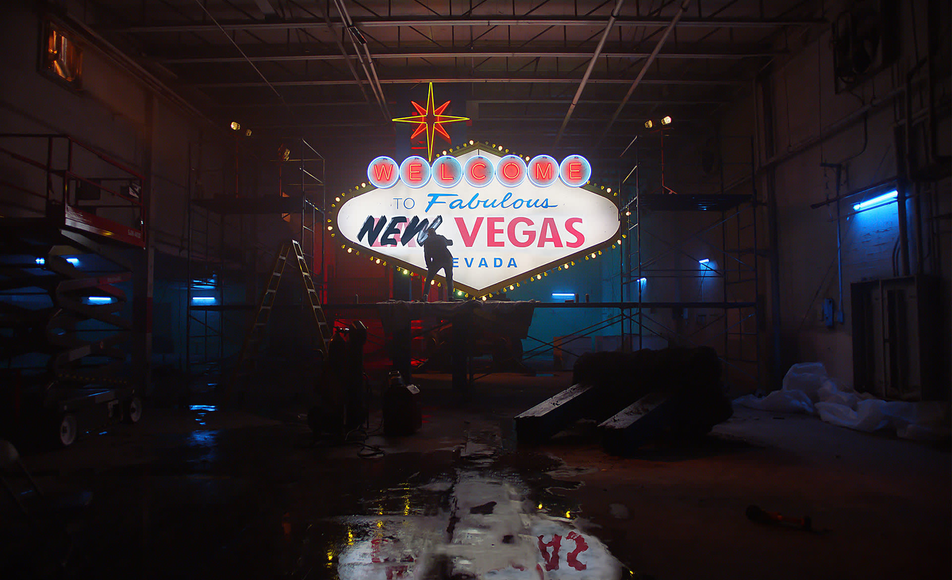 A brightly lit Welcome to Fabulous New Vegas Nevada sign is displayed in a dimly lit industrial warehouse, with scaffolding, ladders, and workers silhouetted around it. The floor reflects the colorful lights.