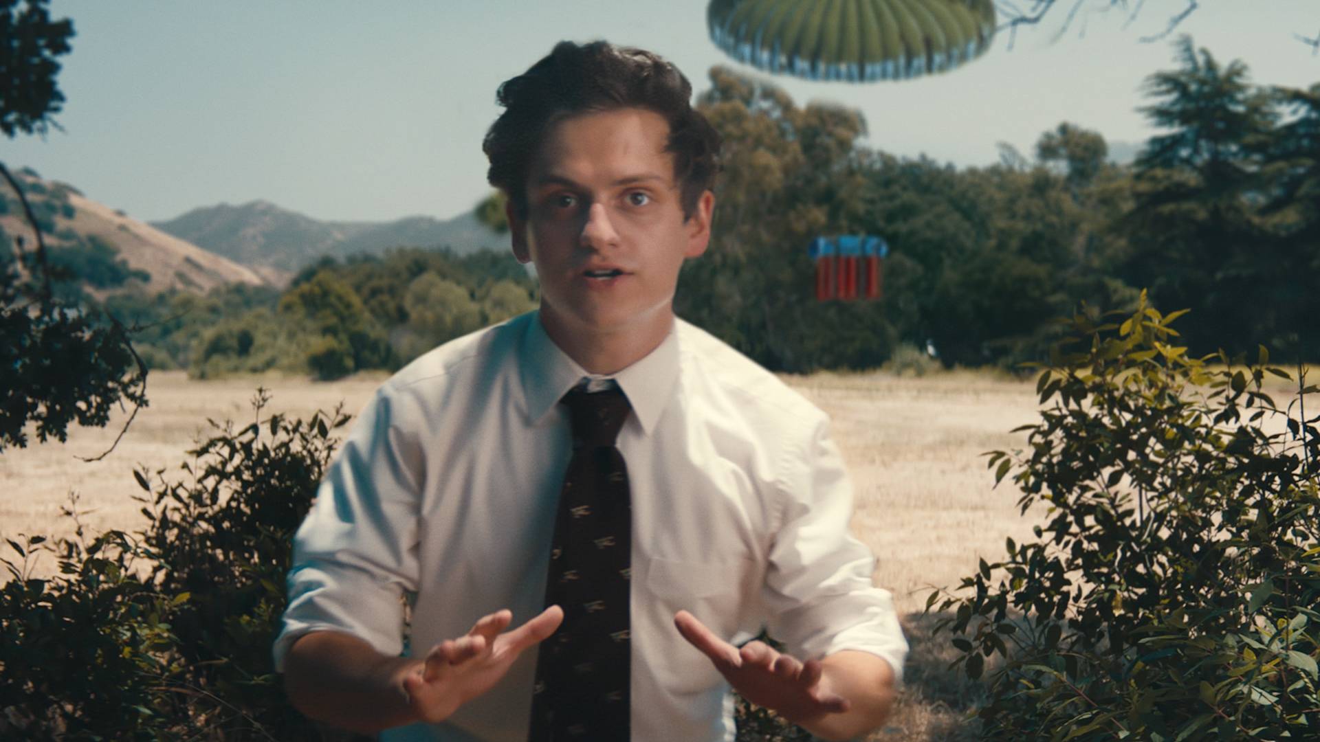 A young man in a white dress shirt and dark tie stands outdoors with his hands raised, as if motioning to calm someone. Behind him, a green parachute and red object are descending from the sky amid trees and hills.