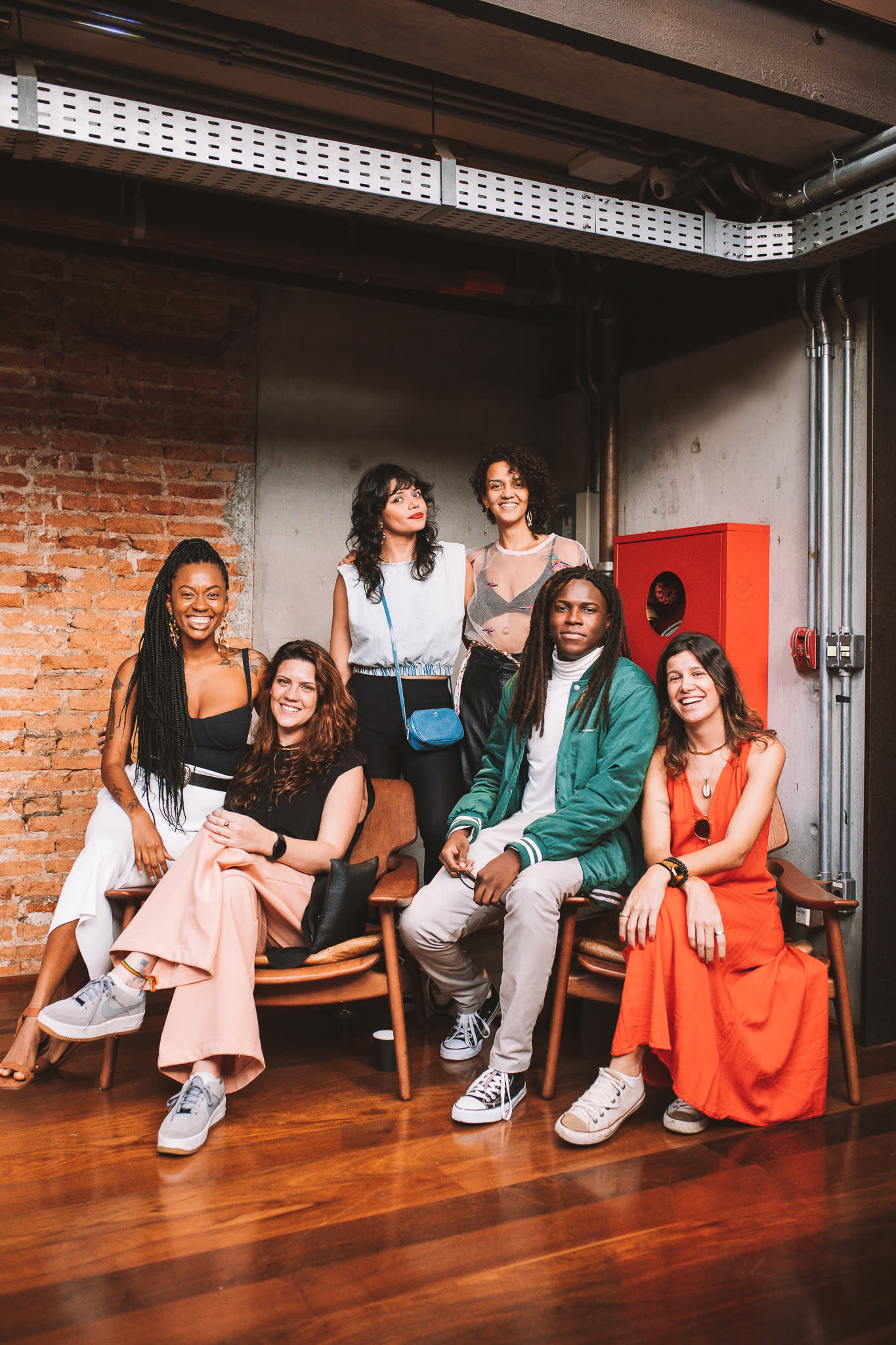 Six people pose and smile together indoors, with four sitting on wooden chairs and two standing behind them. The background features a brick wall and exposed metal pipes. The group appears relaxed and casually dressed.