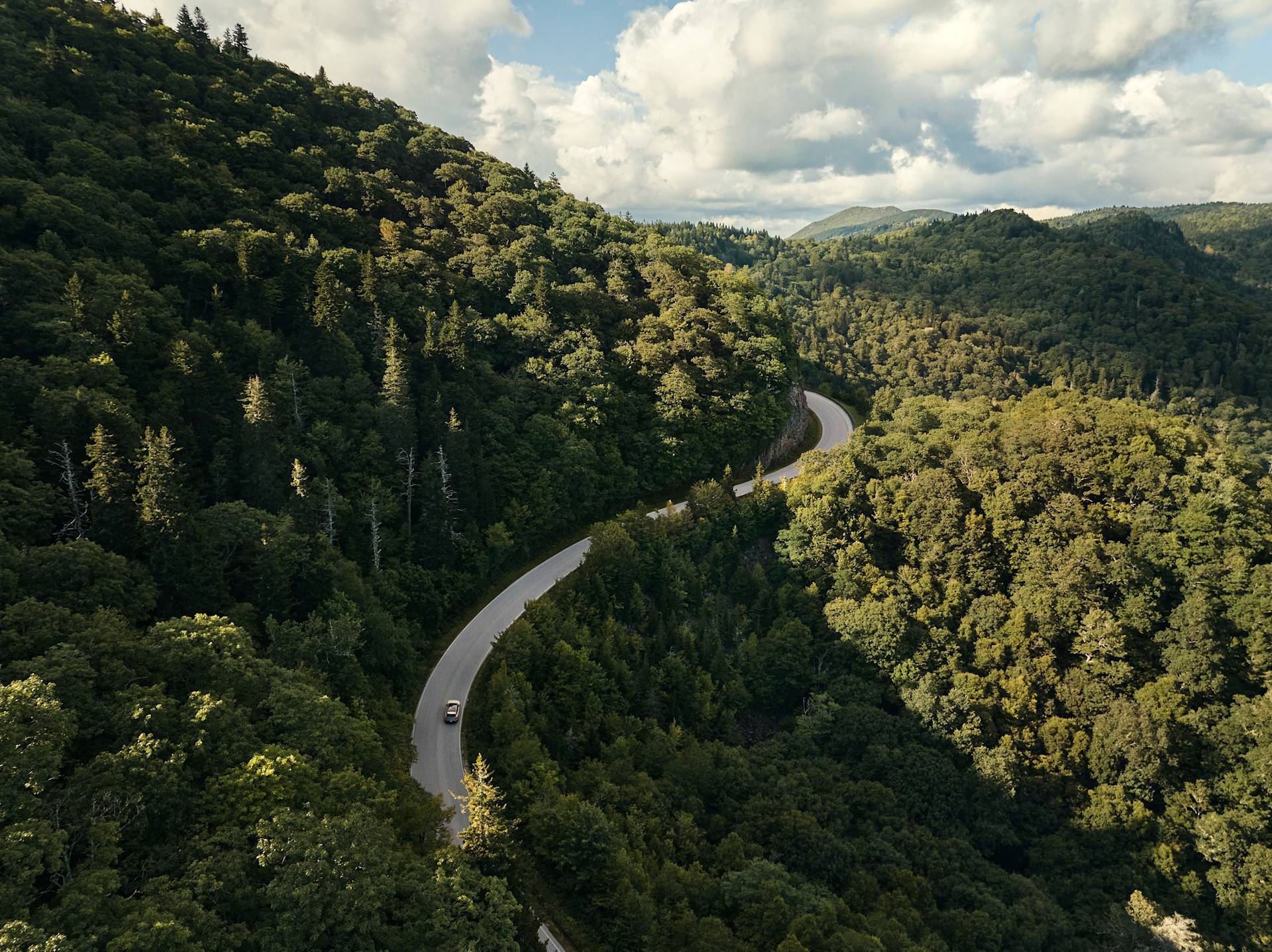 A winding road curves through dense green forested hills under a partly cloudy sky, with a single car driving along the roadway.