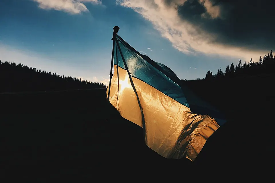 A tattered blue and yellow flag waves in the wind with sunlight shining through it, set against a backdrop of dark hills and a dramatic, cloudy sky at sunset.
