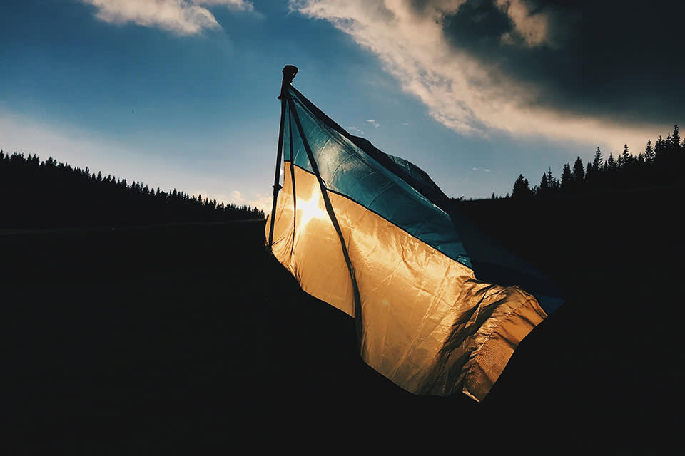 A tattered blue and yellow flag waves in the wind with sunlight shining through it, set against a backdrop of dark hills and a dramatic, cloudy sky at sunset.