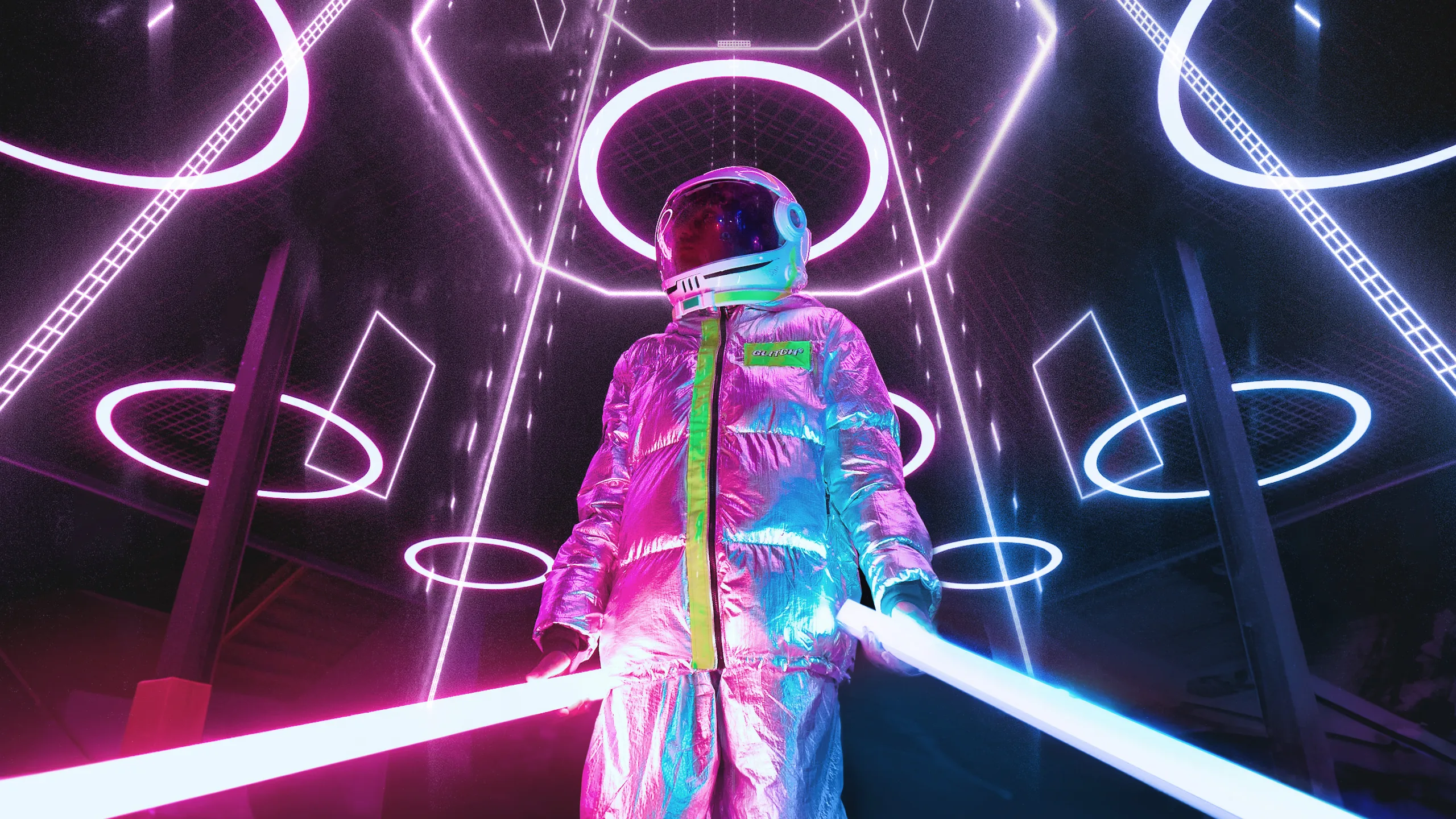 A person in a reflective silver spacesuit and helmet stands in a futuristic room with glowing neon rings and lines, holding two illuminated light sticks, one pink and one blue.