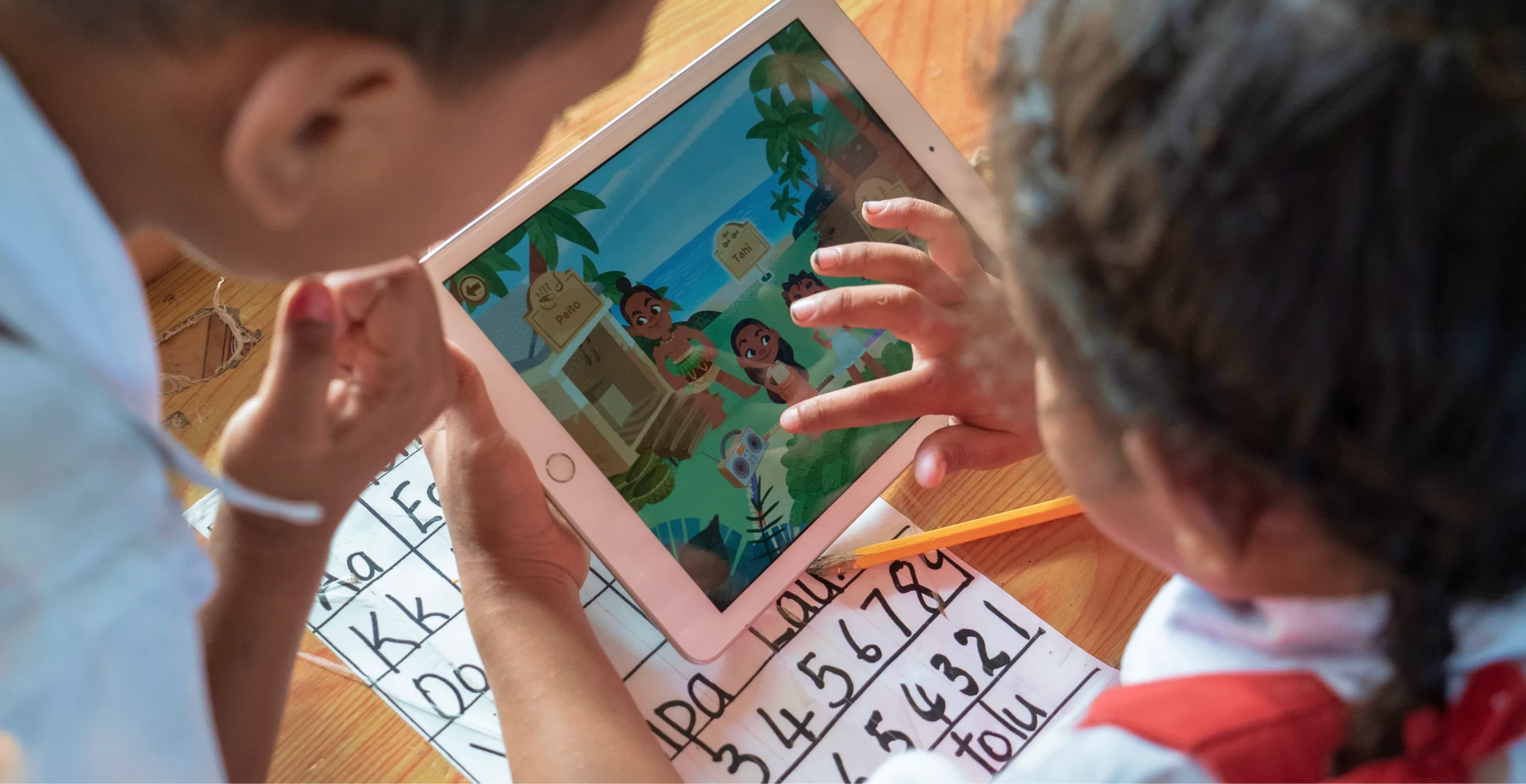 Two children use a tablet displaying a colorful cartoon while sitting at a wooden desk with writing practice sheets and a pencil.