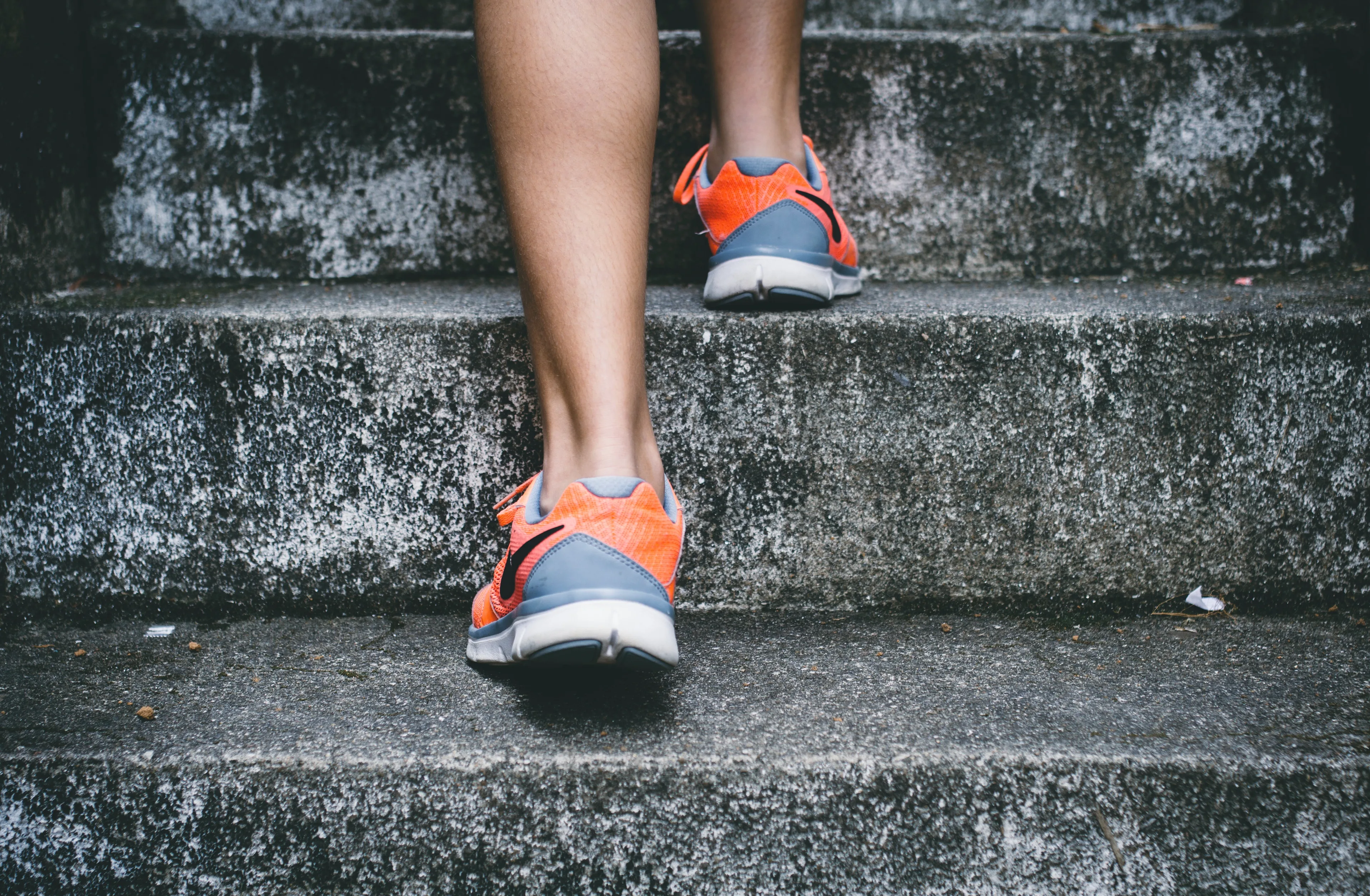 Close-up of a person’s legs as they walk up concrete stairs, wearing orange and gray athletic sneakers. The scene suggests exercise or an active lifestyle.