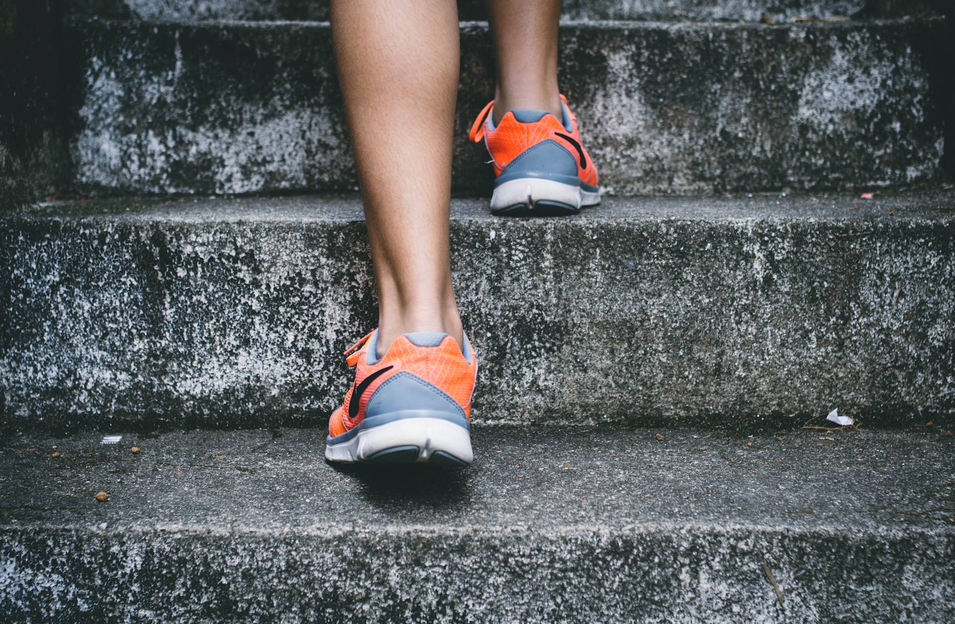 Close-up of a person’s legs as they walk up concrete stairs, wearing orange and gray athletic sneakers. The scene suggests exercise or an active lifestyle.