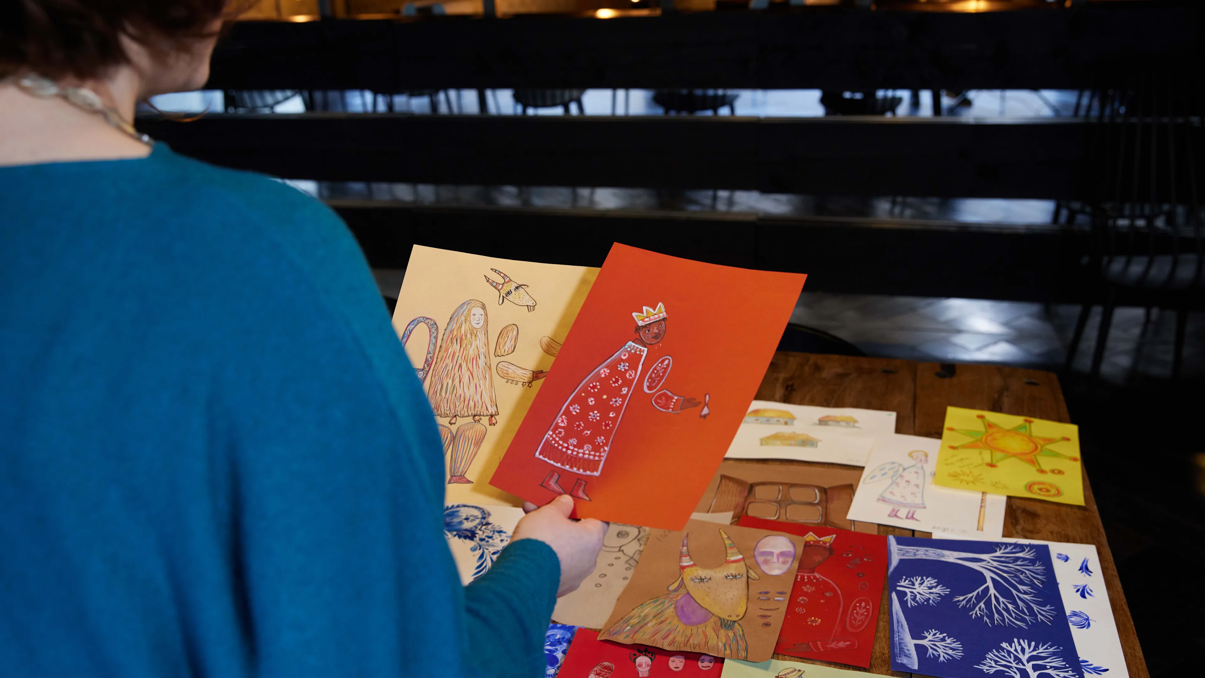 A person in a blue sweater holds a red drawing of a crowned figure over a table covered with colorful, hand-drawn art, including angels, animals, and stars.