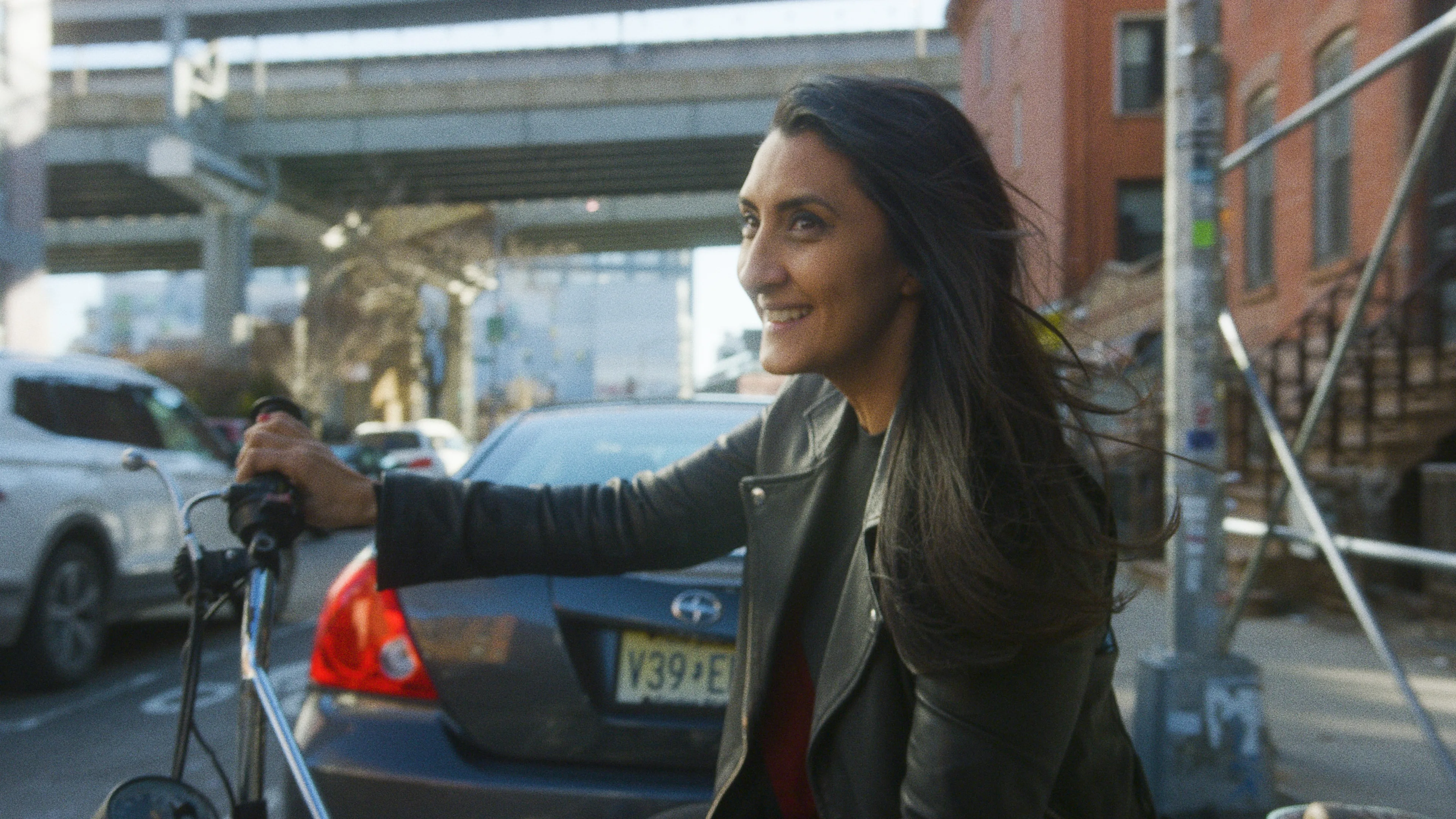 A woman with long dark hair smiles while riding a bicycle on a city street, with cars and an overpass in the background. She is wearing a black jacket and the weather appears to be clear.