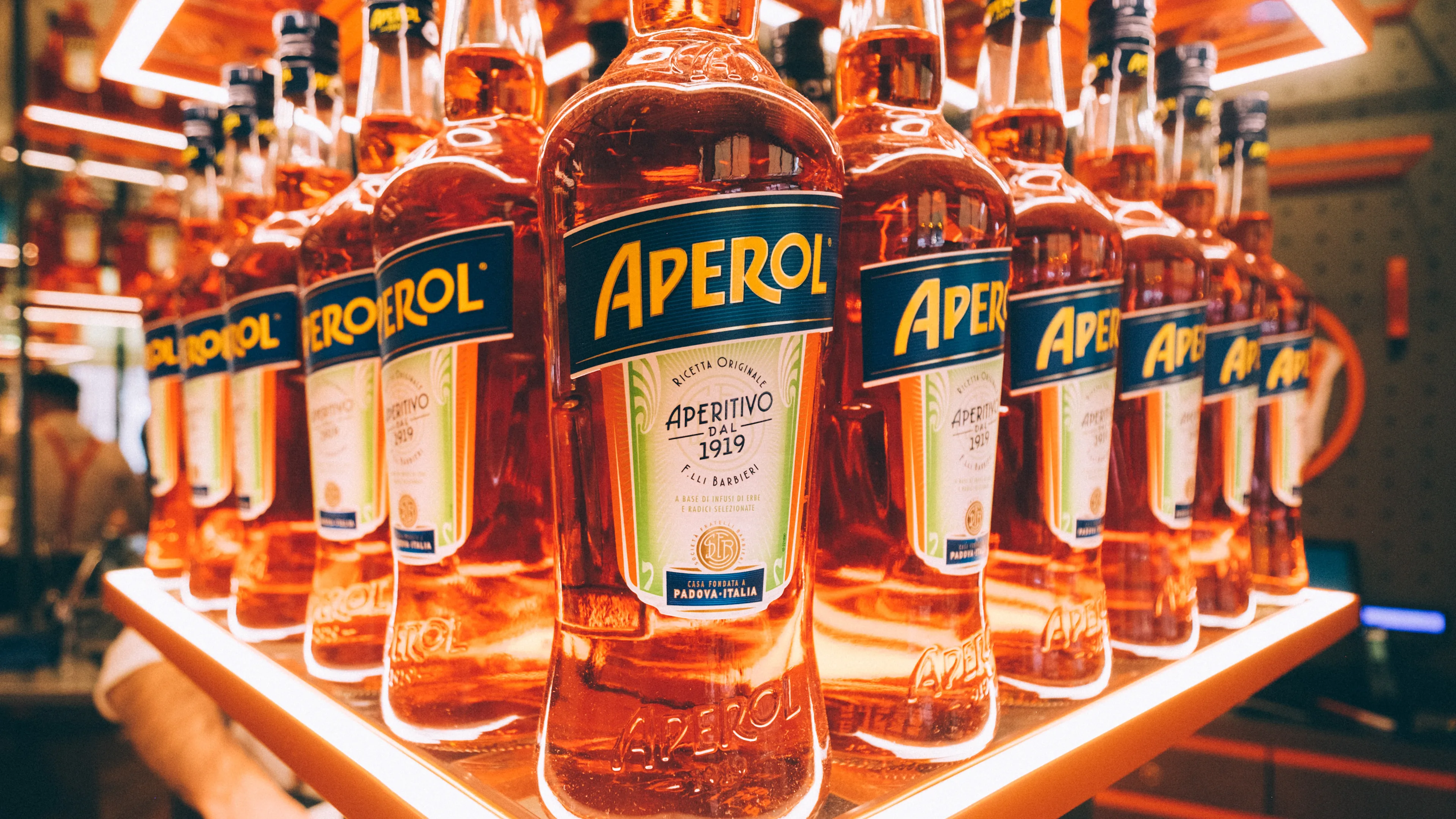 Multiple bottles of Aperol, an orange-colored Italian aperitif, are displayed on illuminated shelves in a bar or store setting. The bottles are arranged in neat rows with prominent blue and yellow labels.