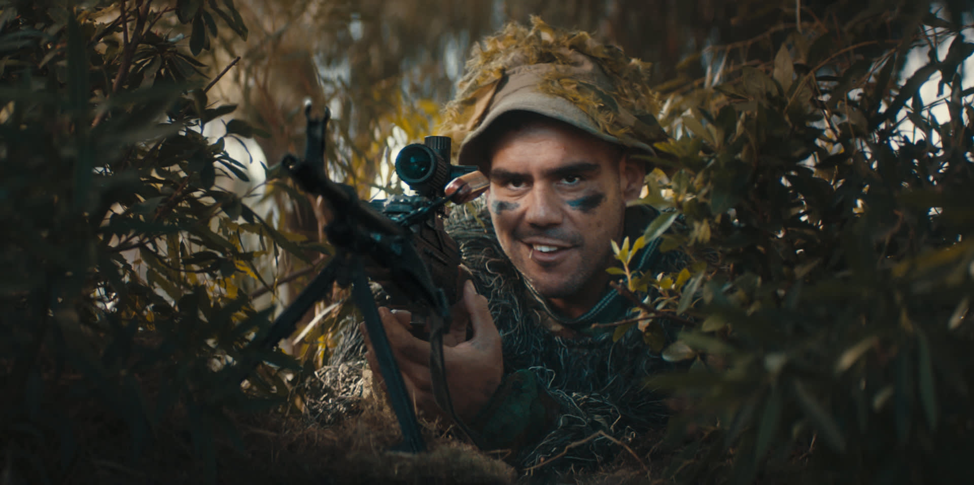 A person in camouflage gear and face paint aims a rifle with a scope while hiding in dense foliage, blending into the surroundings.