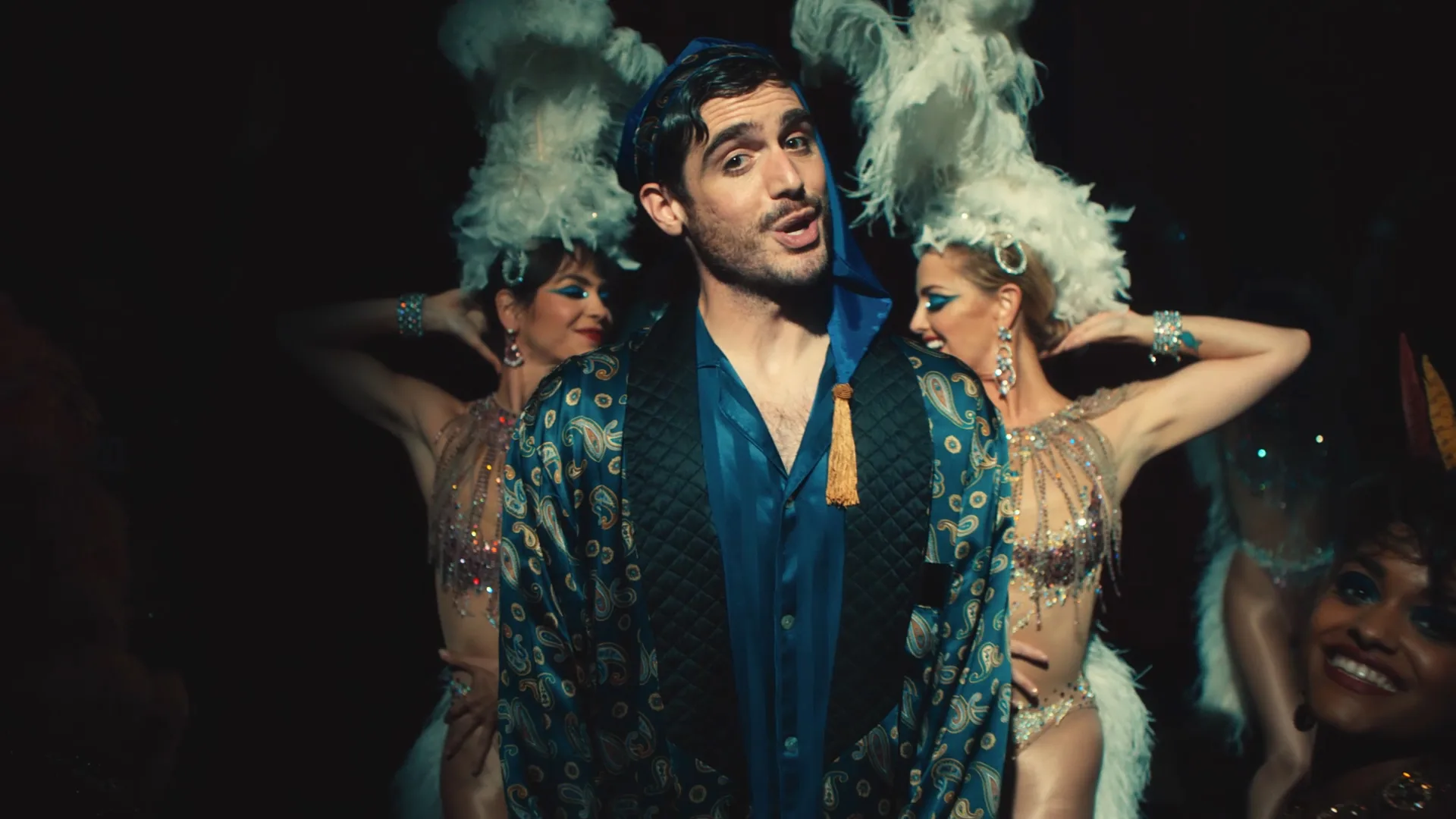 A man in a patterned robe and blue headscarf stands center stage, flanked by two women in elaborate feathered headdresses and sparkling costumes, performing against a dark background.