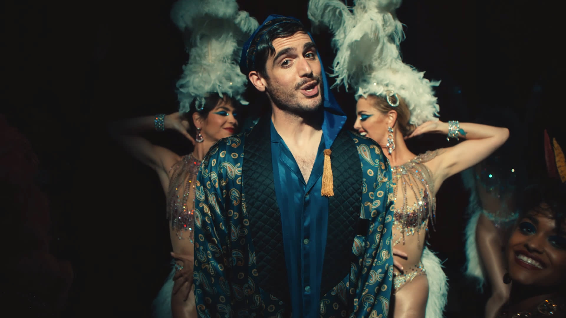 A man in a patterned robe and blue headscarf stands center stage, flanked by two women in elaborate feathered headdresses and sparkling costumes, performing against a dark background.