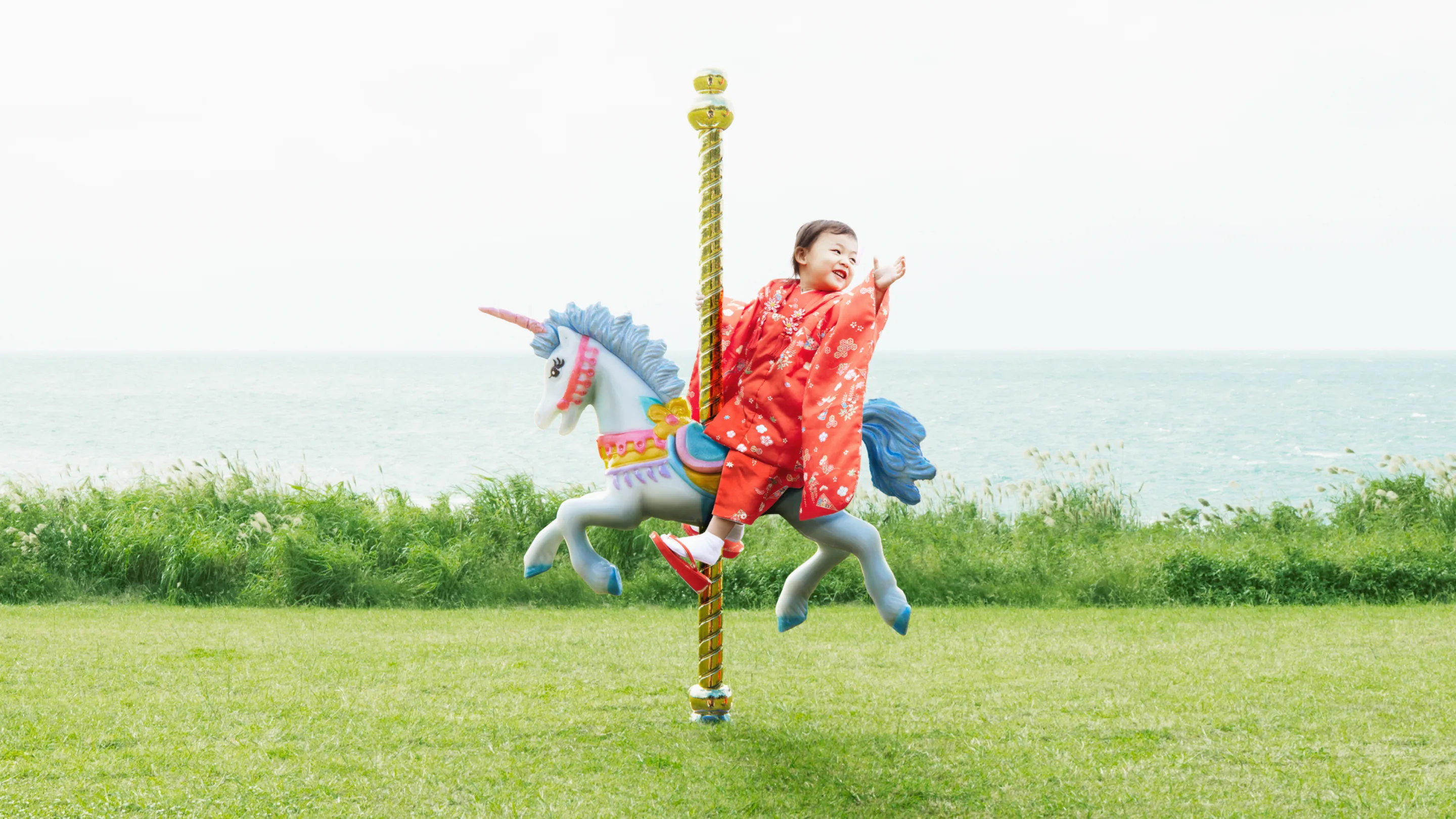 A child in a bright red outfit joyfully rides a colorful carousel unicorn outdoors on green grass, with the ocean and blue sky in the background.