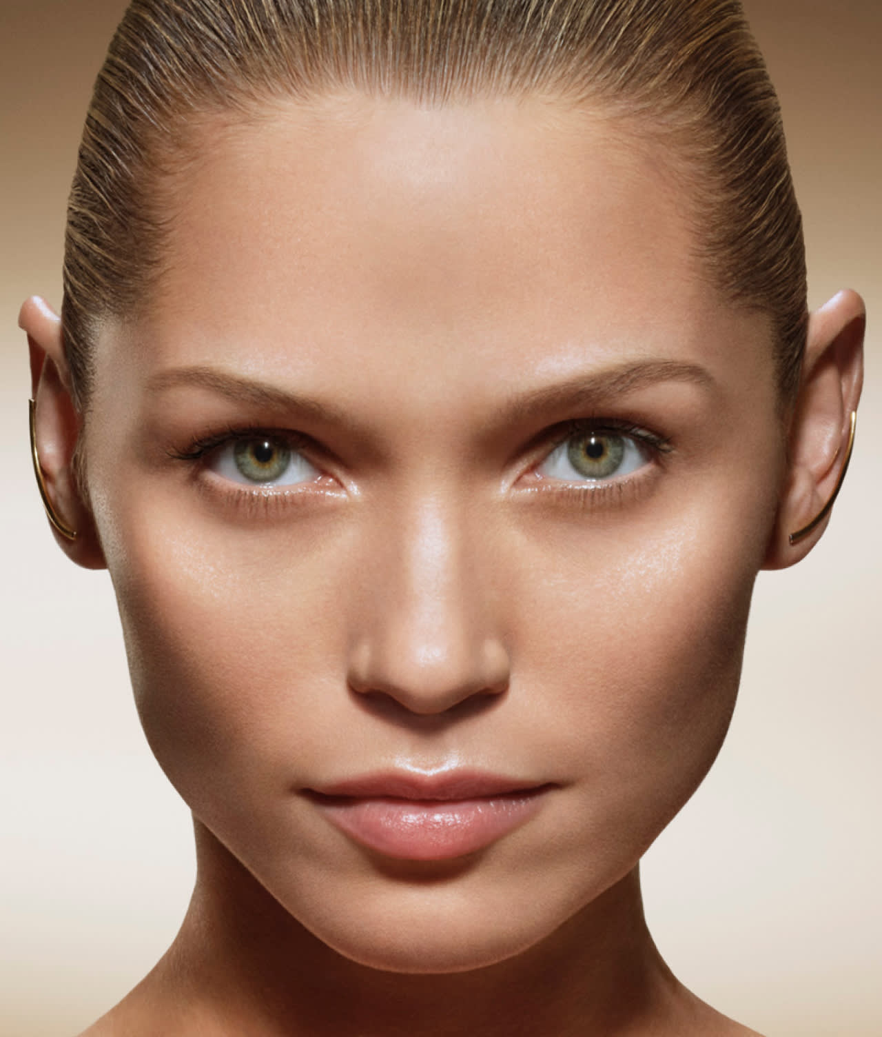 Close-up of a person with slicked-back hair, green eyes, clear skin, and natural makeup, looking directly at the camera against a neutral background.