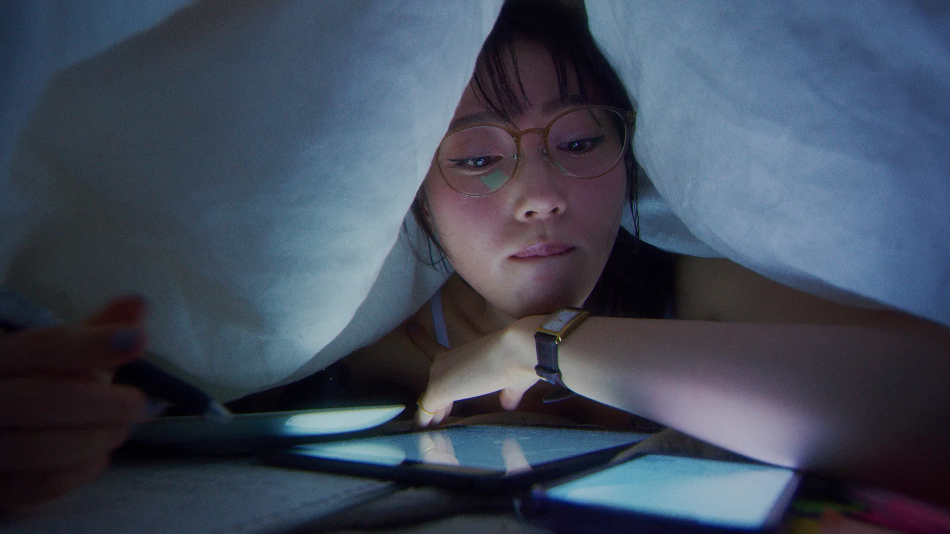 A young woman wearing glasses lies under a blanket, surrounded by glowing screens from multiple devices, concentrating as she uses a pen and looks at a tablet or phone.