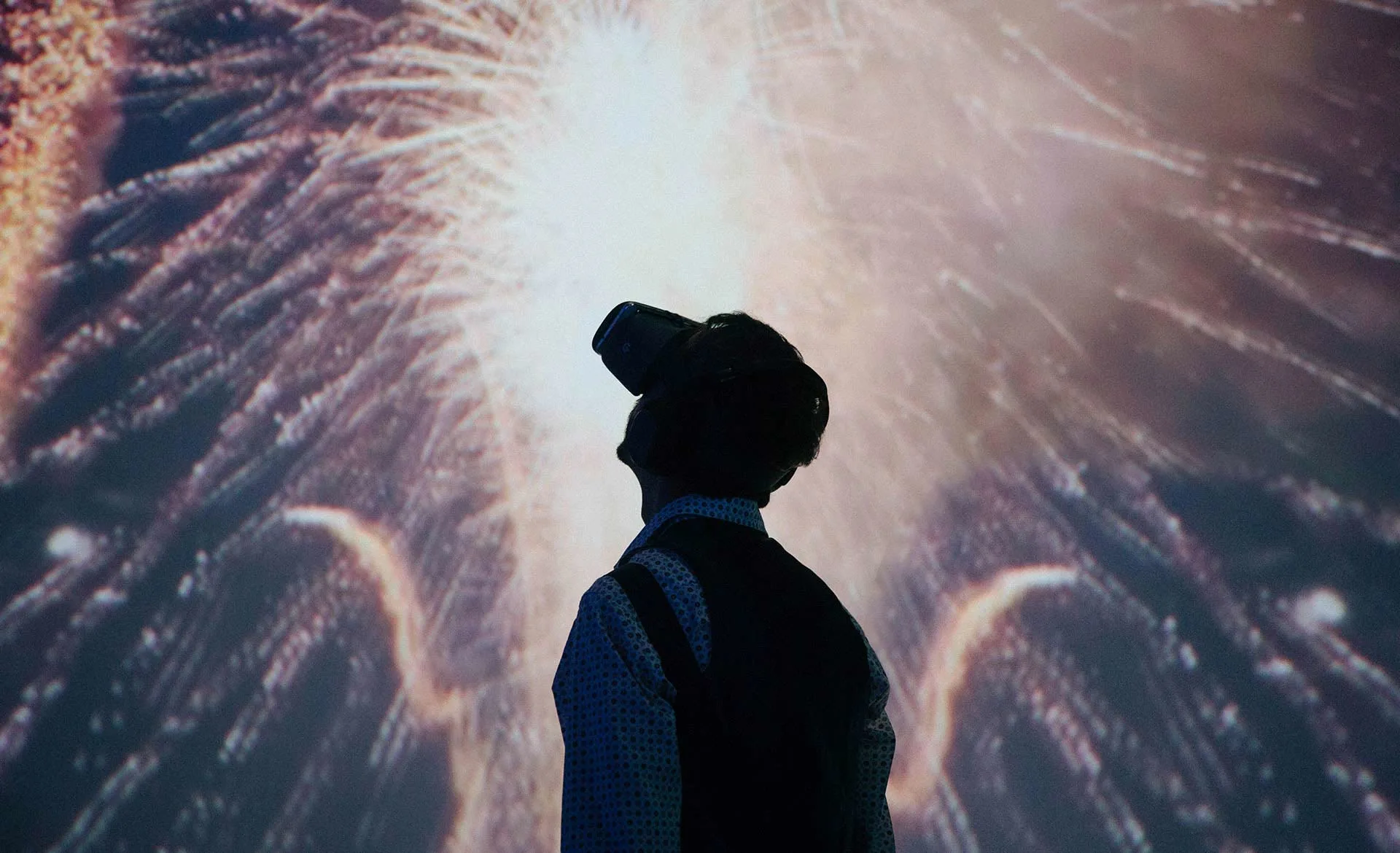 A person wearing a virtual reality headset stands in front of a vibrant, illuminated background resembling exploding fireworks.