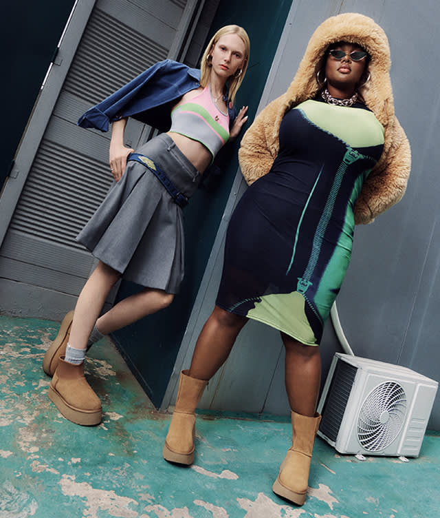 Two women pose confidently in stylish outfits and tan boots. One wears a grey skirt, crop top, denim jacket, and the other wears a fitted dress with a furry hood and sunglasses. They stand on a weathered floor near an AC unit.