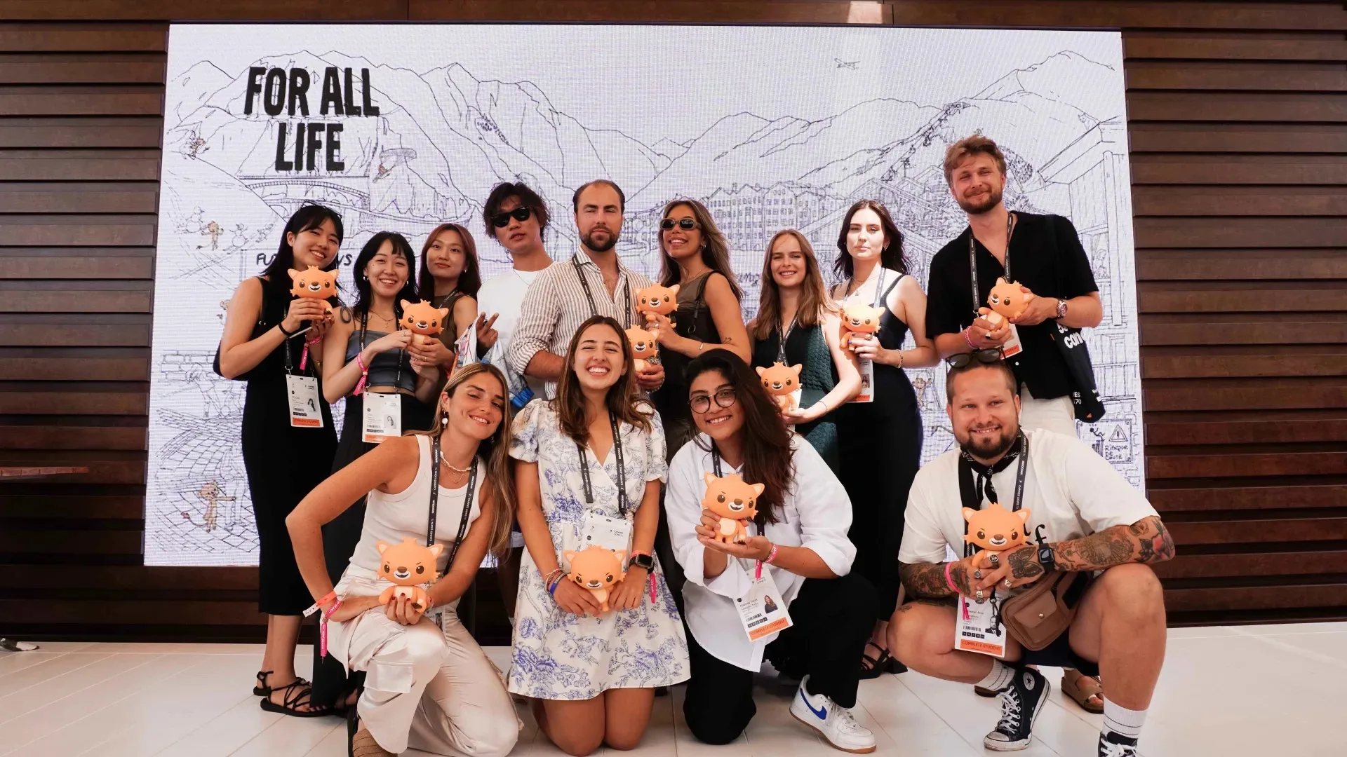 A group of people pose and smile in front of a mountain-themed backdrop that reads “FOR ALL LIFE.” Most hold small orange cat plush toys and wear event badges. They are dressed in casual summer clothing.