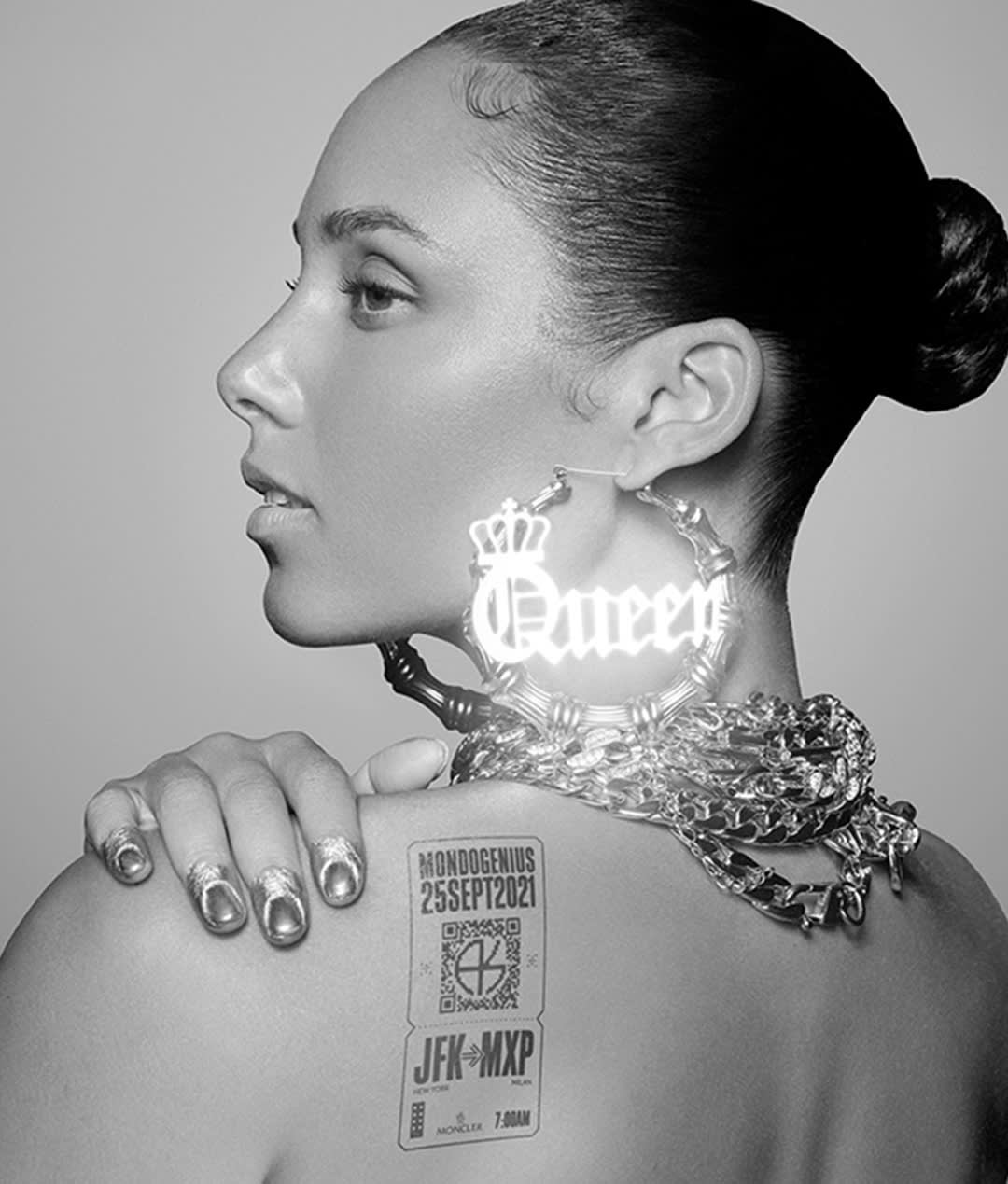 A woman in profile with her hair in a sleek bun, wearing large Queen hoop earrings and thick chains. She has metallic silver nail polish and a ticket-like tattoo on her shoulder with MONOGENIUS 25SEPT2021 visible.