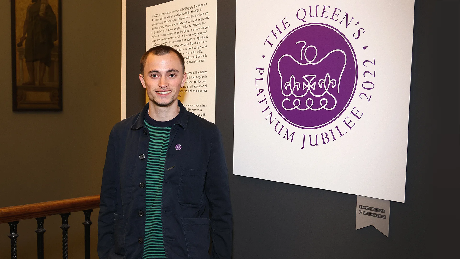 A young man stands smiling next to a sign that reads The Queens Platinum Jubilee 2022 with a stylized crown emblem, inside an indoor exhibition space.