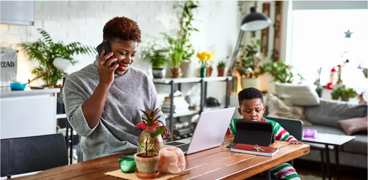 A woman smiles while talking on the phone and working on a laptop at a table, next to a young child using a tablet. The room is bright, filled with houseplants and decorated for the holidays.