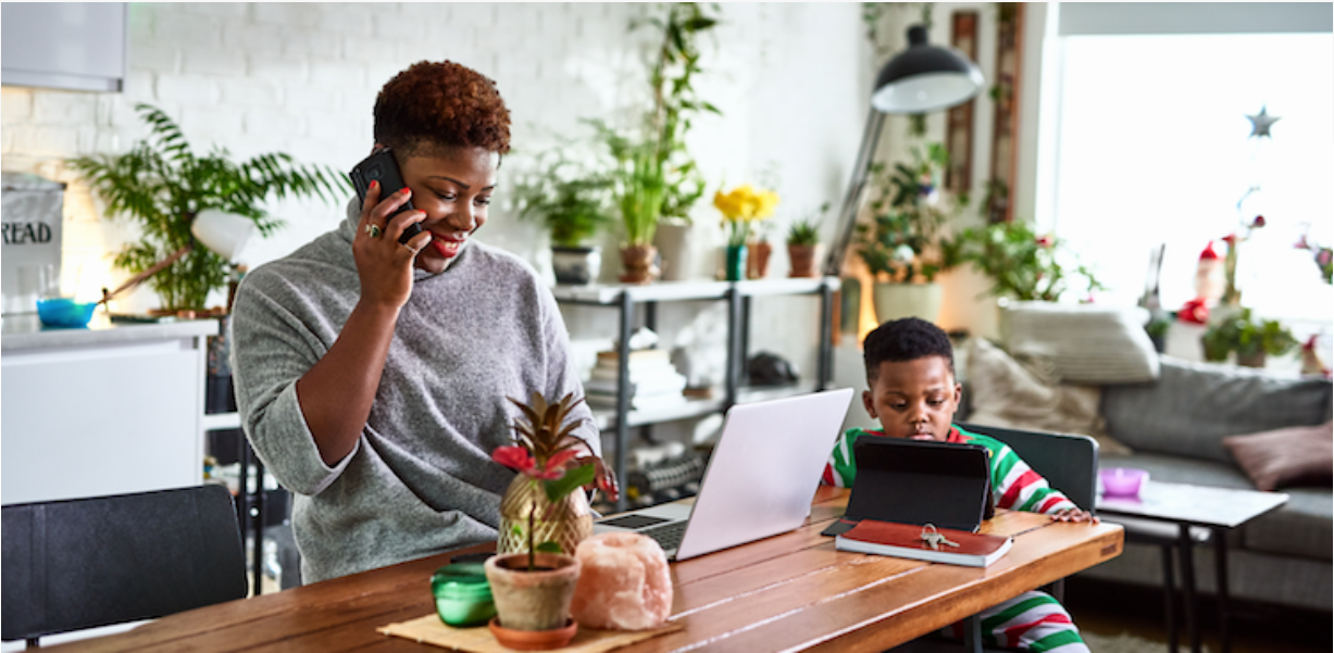 A woman smiles while talking on the phone and working on a laptop at a table, next to a young child using a tablet. The room is bright, filled with houseplants and decorated for the holidays.
