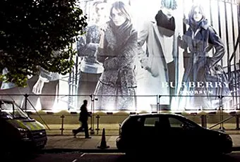 A person walks past a large illuminated Burberry fashion billboard at night, featuring models in stylish clothing. Cars are parked on the street in front, and part of a tree is visible on the left side.