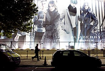 A person walks past a large illuminated Burberry fashion billboard at night, featuring models in stylish clothing. Cars are parked on the street in front, and part of a tree is visible on the left side.