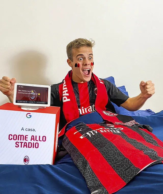 A young soccer fan with face paint and an AC Milan scarf cheers excitedly on a bed, with an AC Milan jersey and a box reading A casa, COME ALLO STADIO nearby, watching a match on a smartphone.