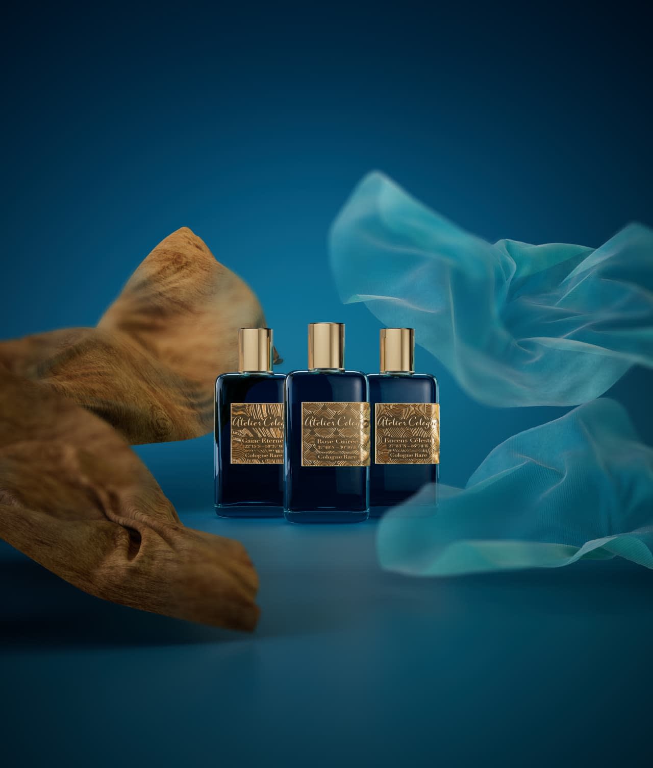 Three elegant blue perfume bottles with gold caps and labels are arranged side by side against a blue background, accented by flowing turquoise fabric and textured brown driftwood decor.